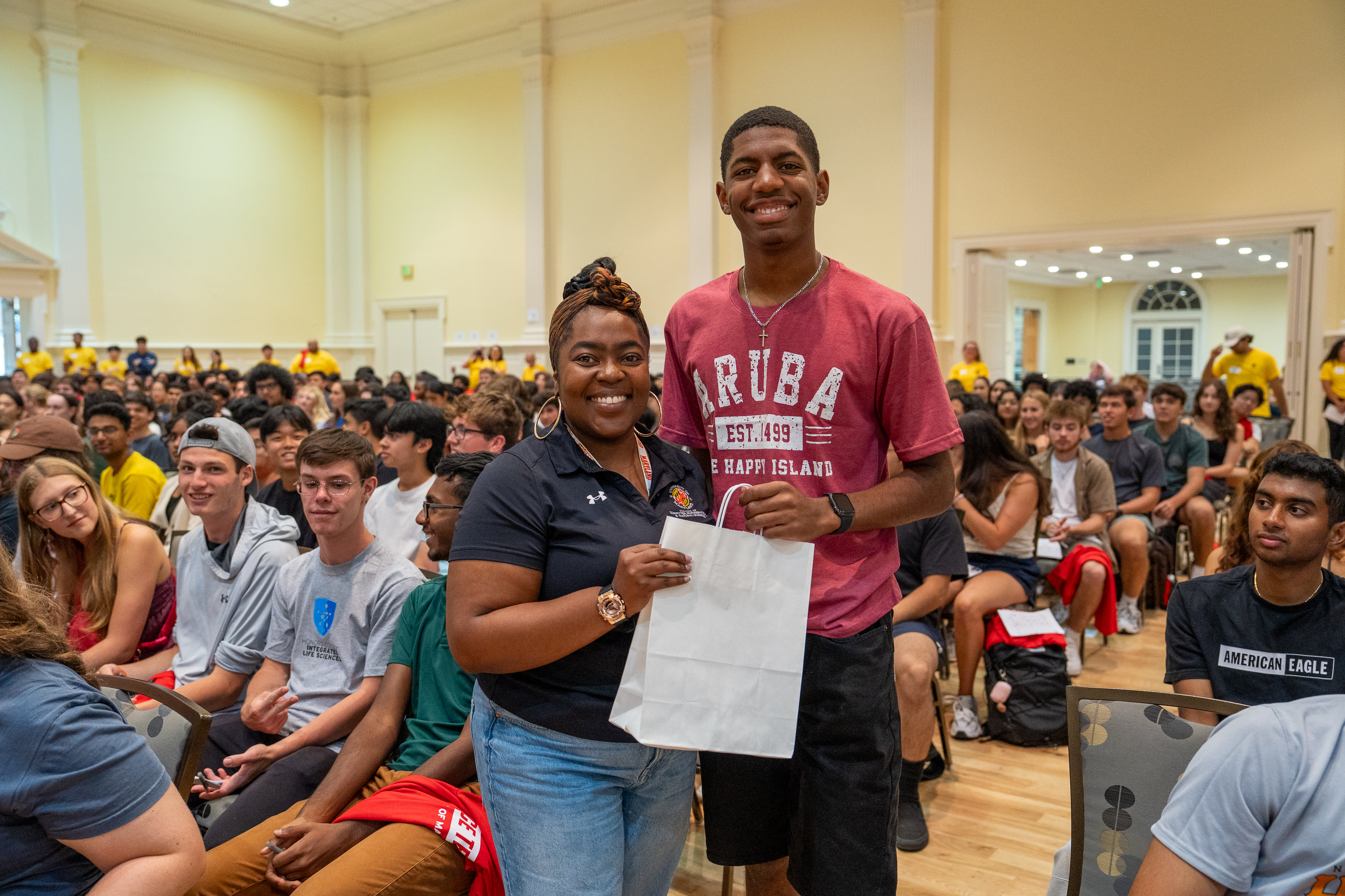 A woman stands with a student holding a paper bag, with an audience seated behind them