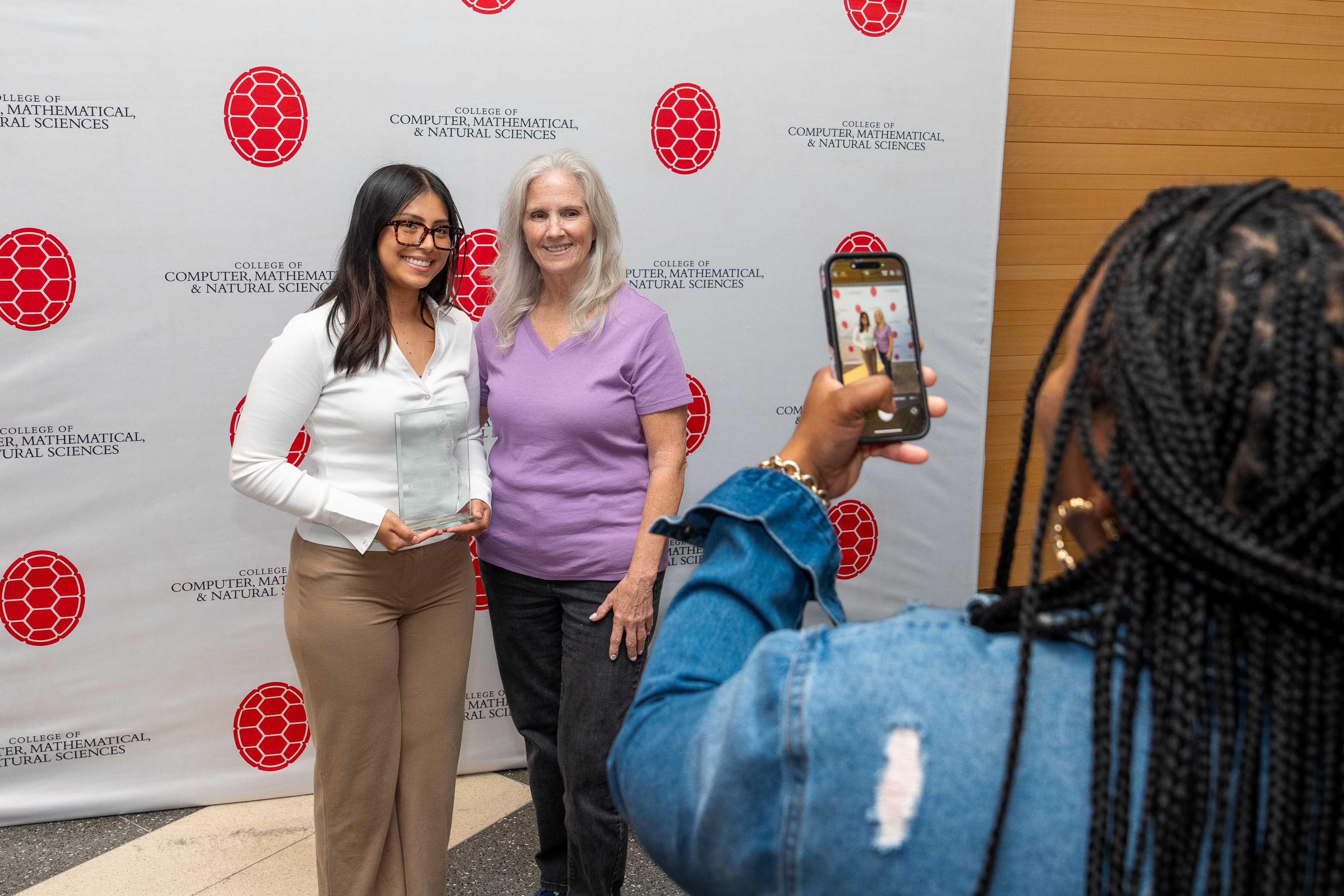 A woman takes a photo on her phone of two people standing in front of a step-and-repeat banner