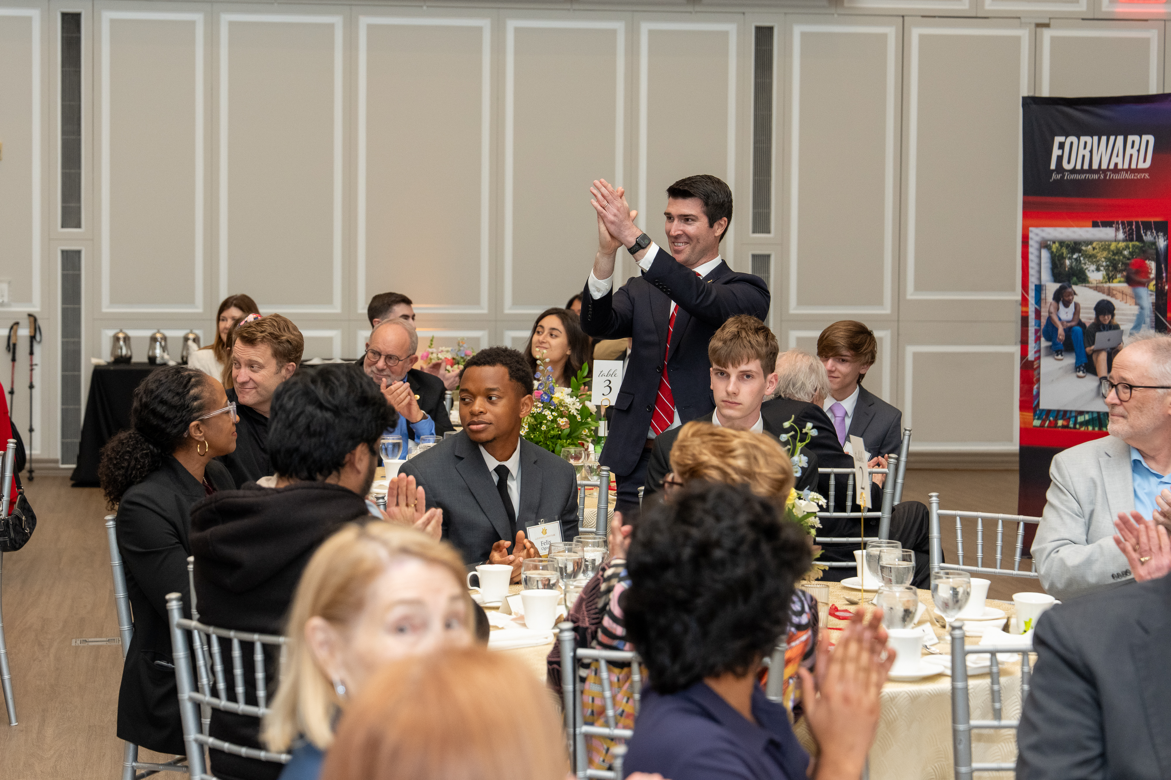 A man wearing a suit claps while surrounded by people seated at tables