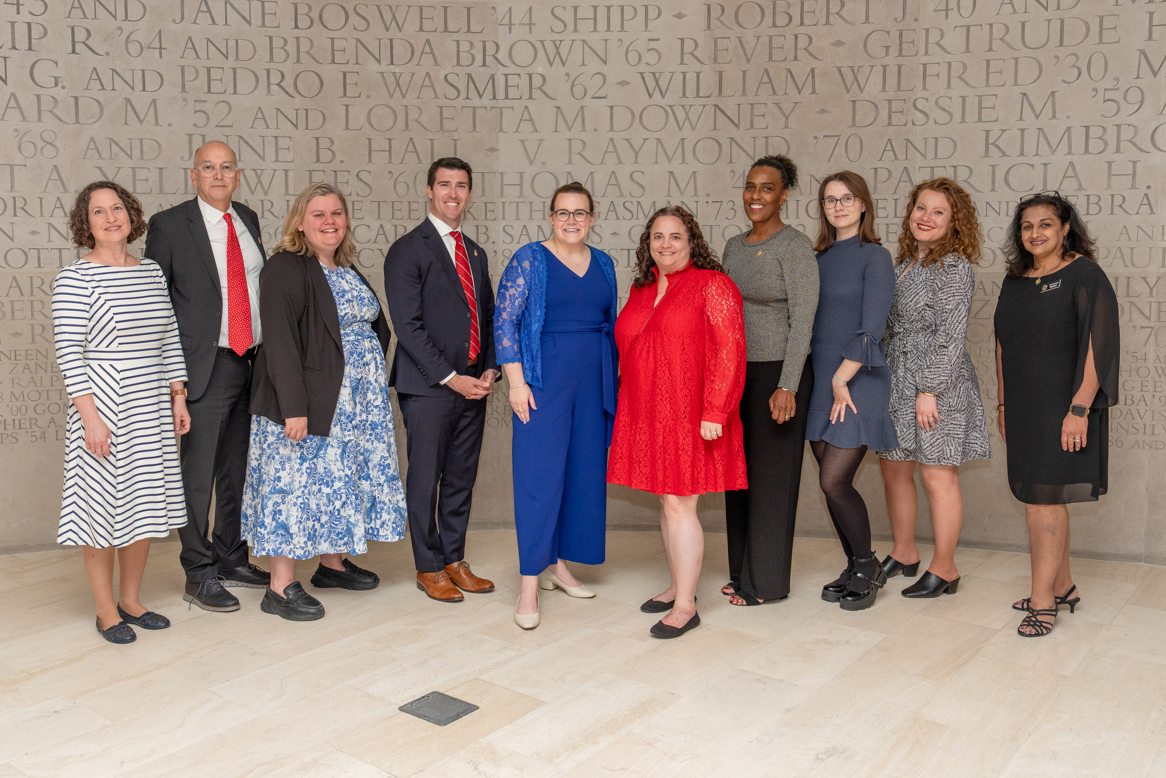 A group of people in semi-formal dress standing in front of a gray wall with engraved text on it