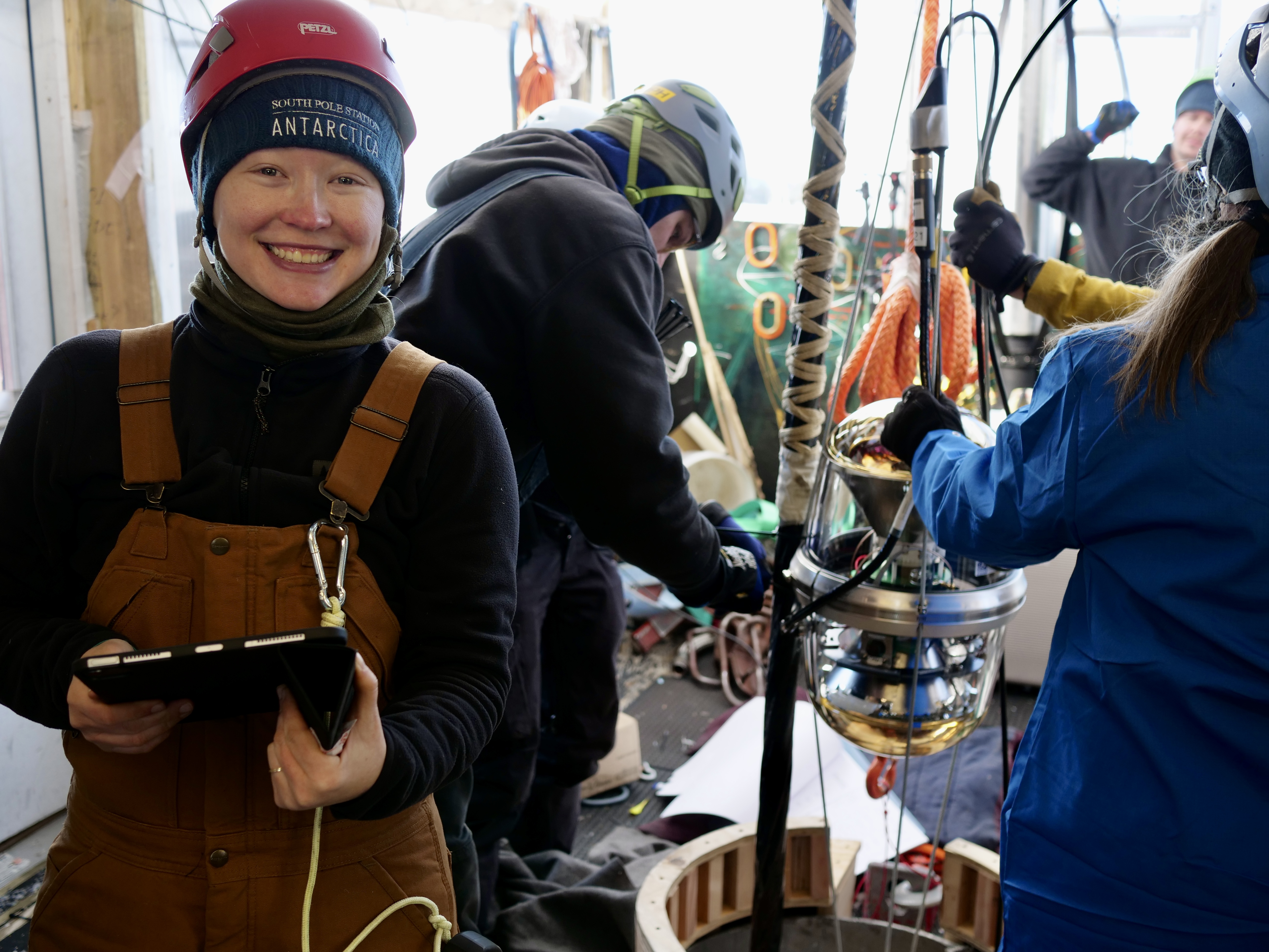 Rachel Procter-Murphy holding an iPad and wearing a hardhat and overalls among people installing equipment at the IceCube observatory.