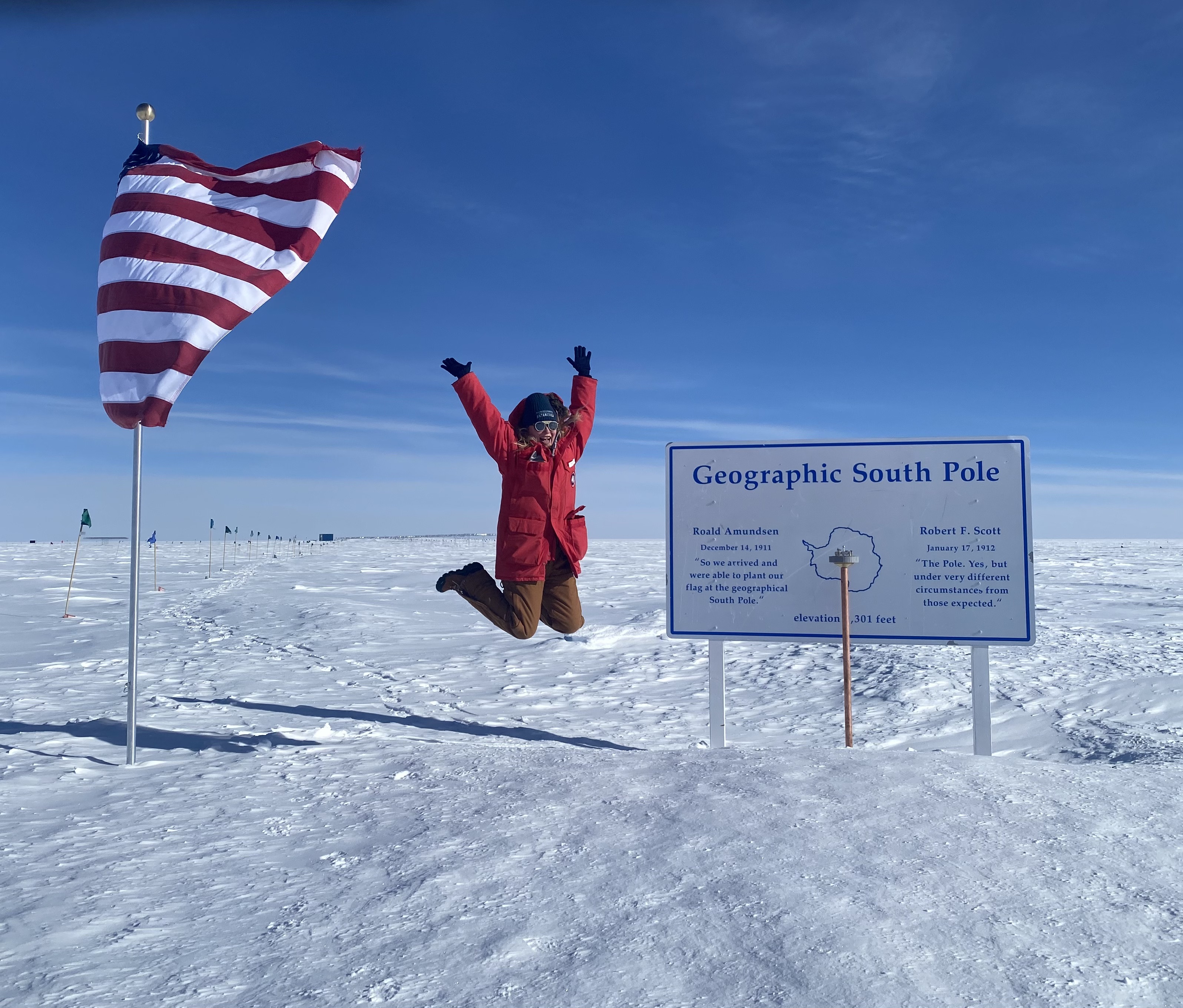 Rachel Procter-Murphy on the ice in a red coat leaping next to an American flag and a sign that reads "Geographic South Pole." 