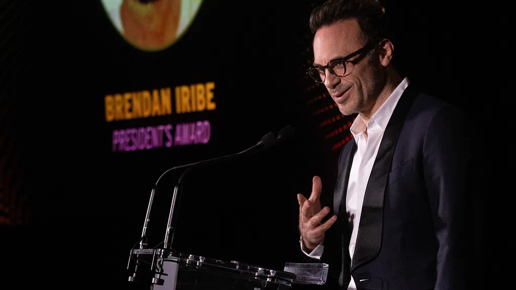 Smiling man speaks at podium. Projected  in the background is his name and the name of the award he is receiving.