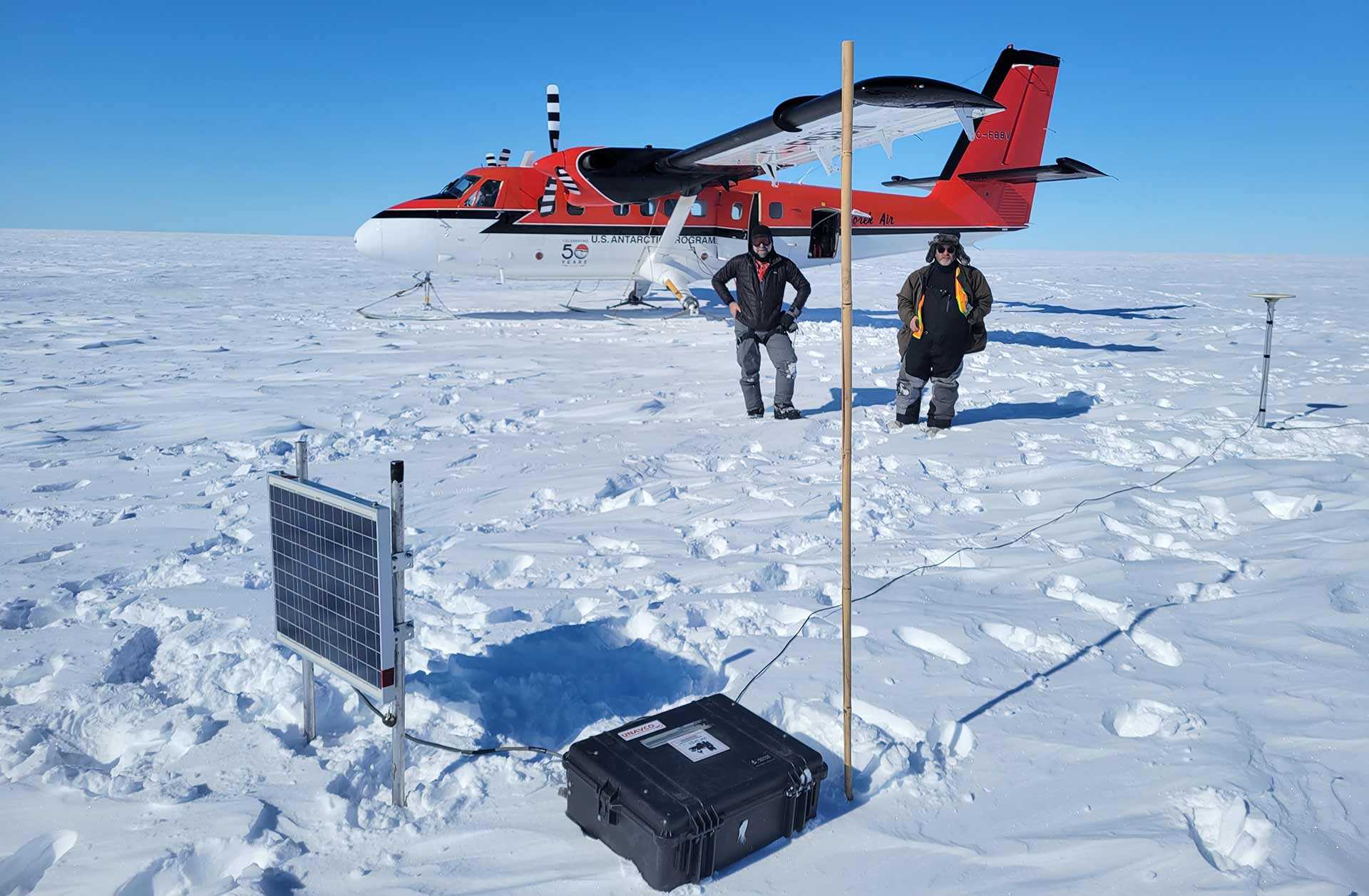 Associate Professor Nick Schmerr, left, with pilot Troy McKerral, behind a GPS station with a solar panel, recording unit, and antenna. The bamboo pole helps the retrieval team find the equipment, since the ice shelf can move a meter per day.