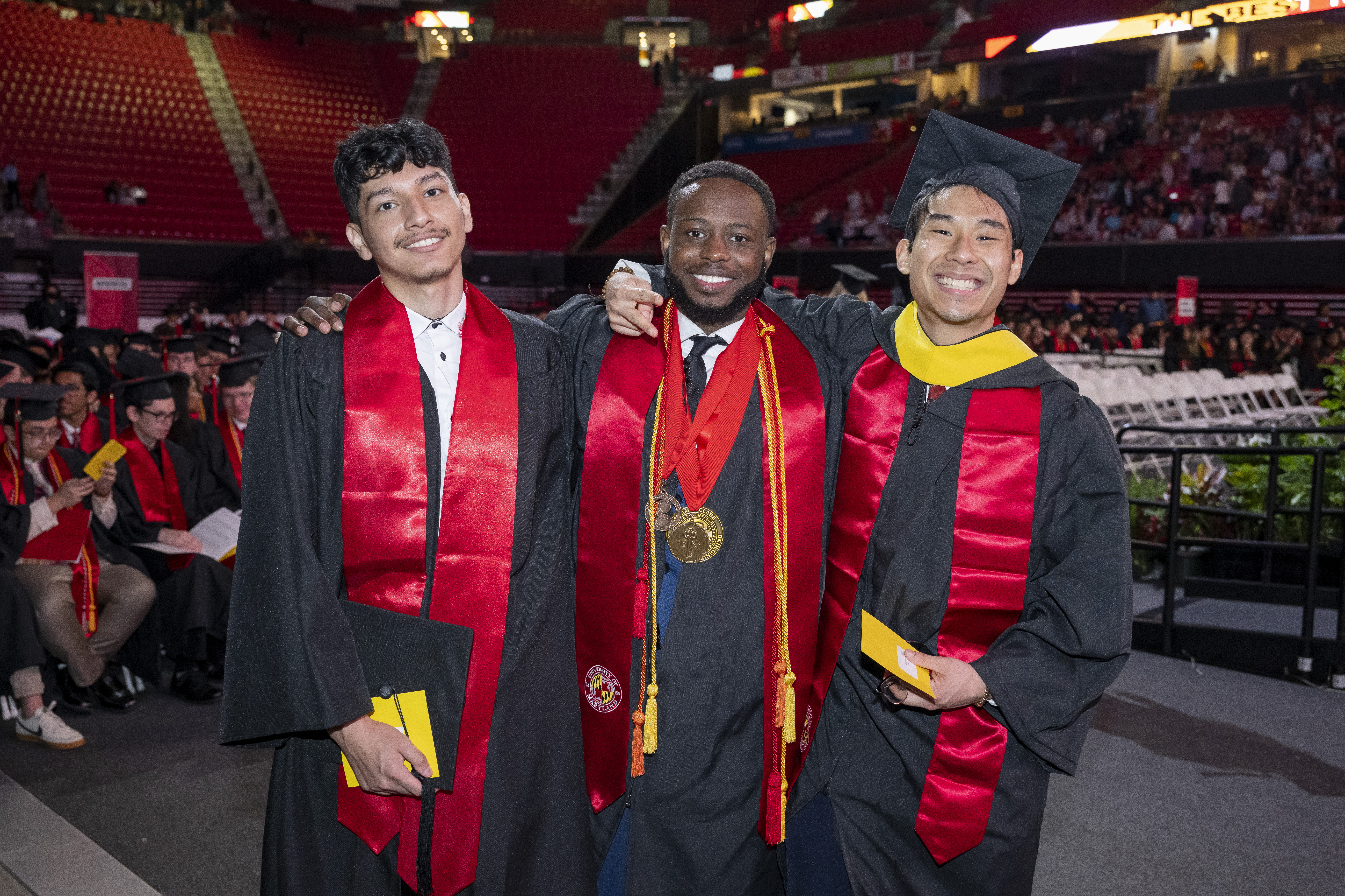 Three students smiling in full regalia before commencement in the Xfinity Center