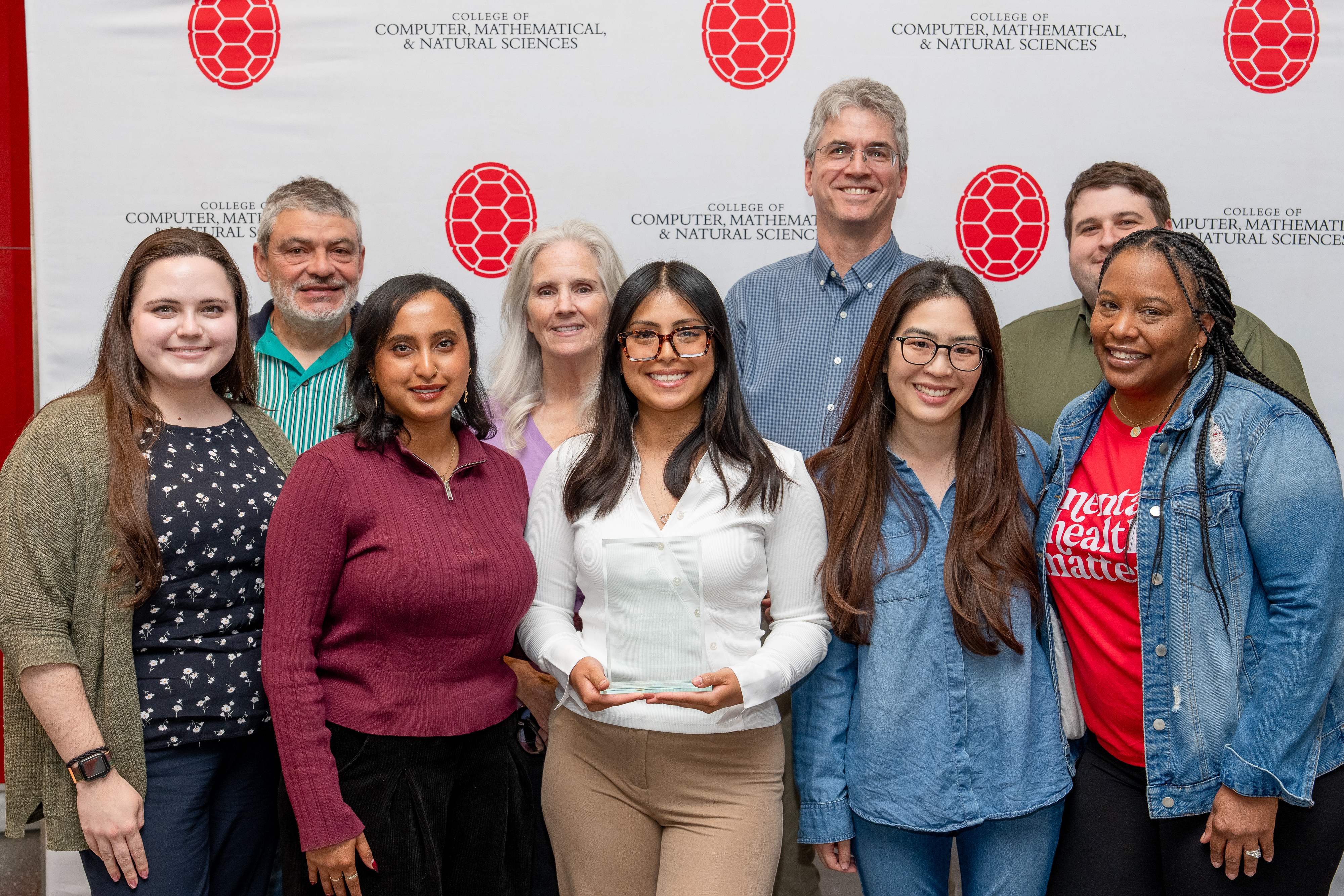 Person holding an award in front of a step-and-repeat banner, surrounded by a group of other people