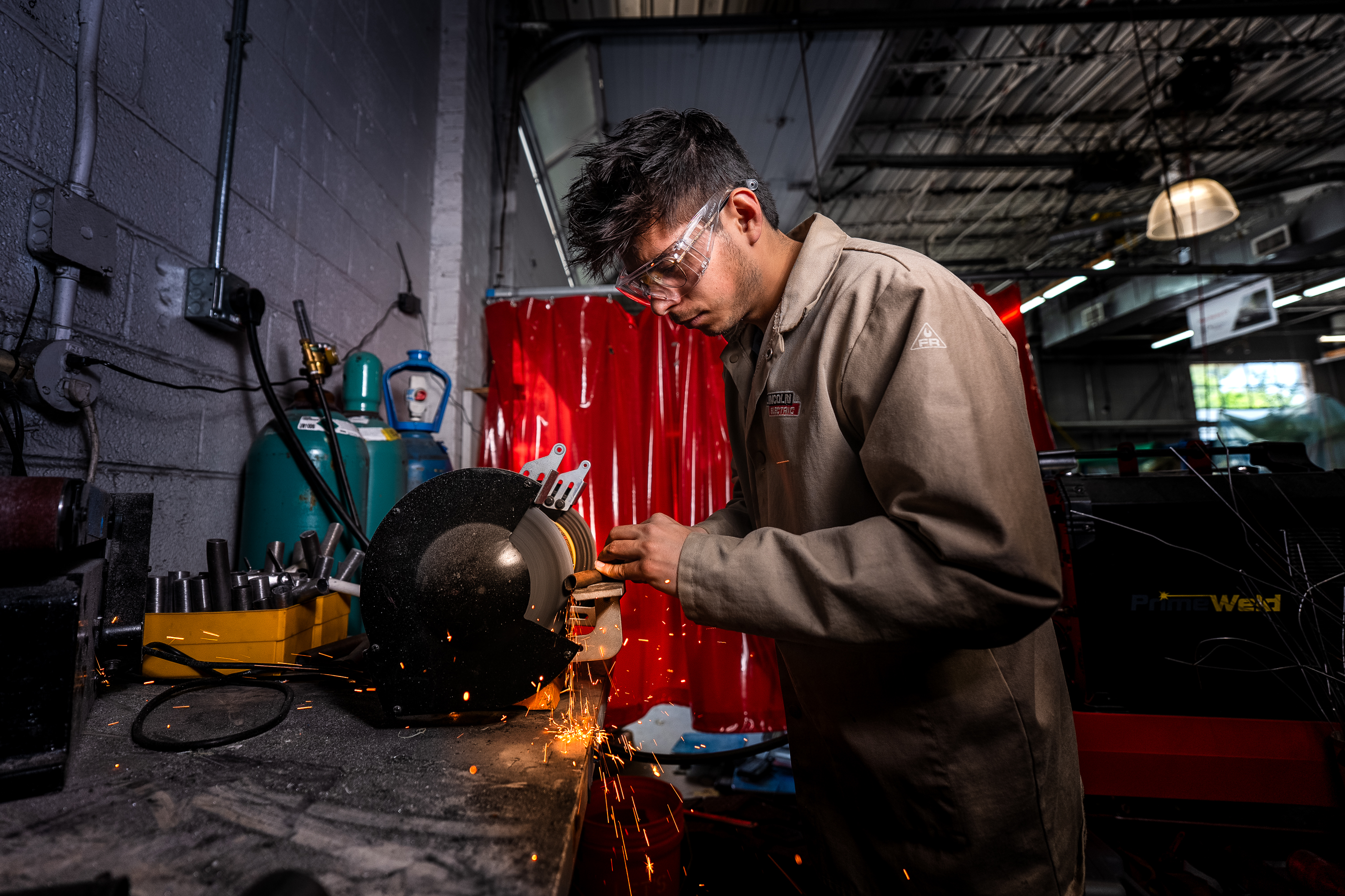 A student wearing safety goggles and a work jacket uses an angle grinder on a piece of metal, sending spars across a workshop bench.