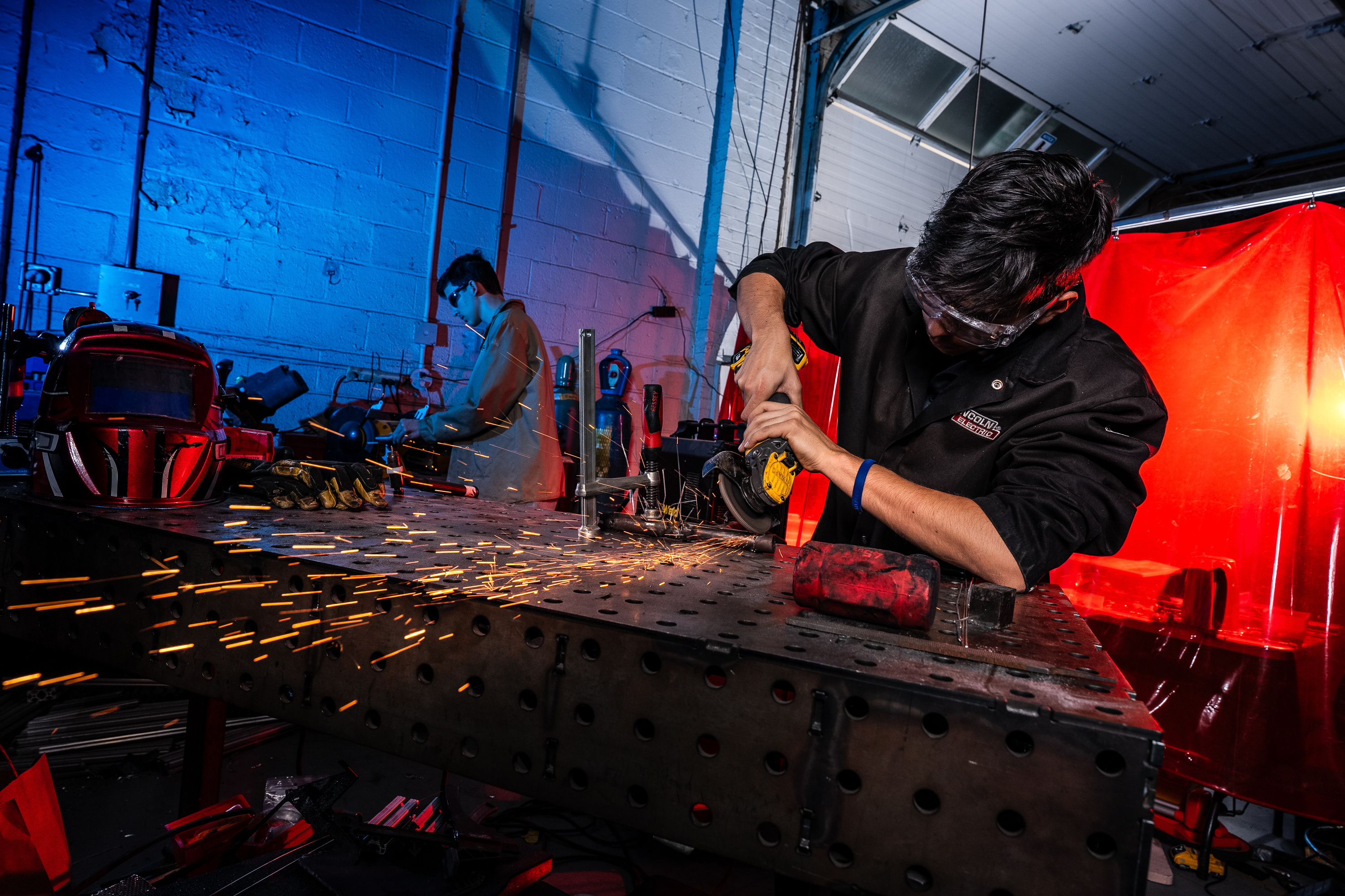 Two students work in a dim car shop—one in the front using an angle grinder, while the other is in the background in blue lighting.