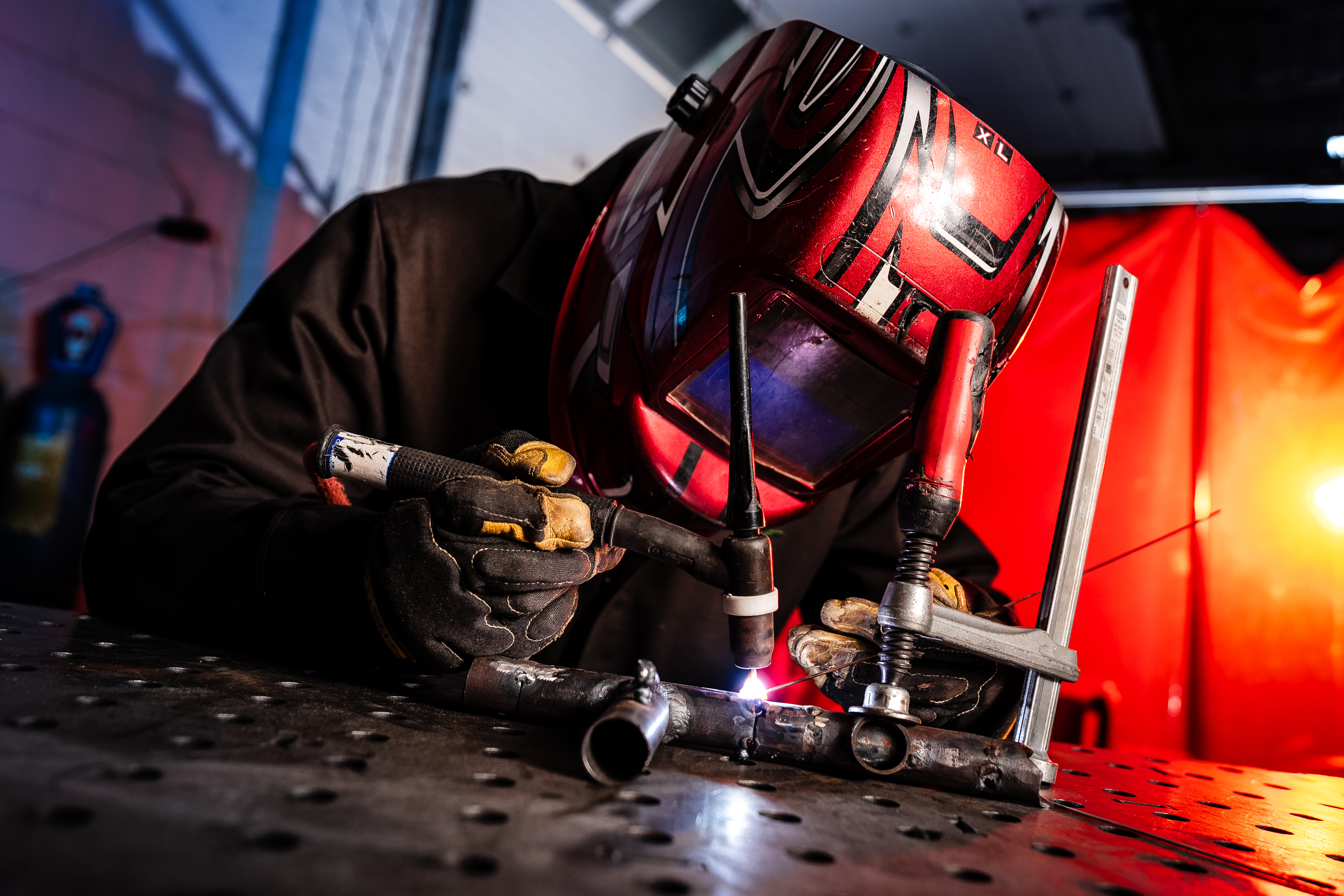 A student in a red welding helmet and leather gloves welds on a table. 