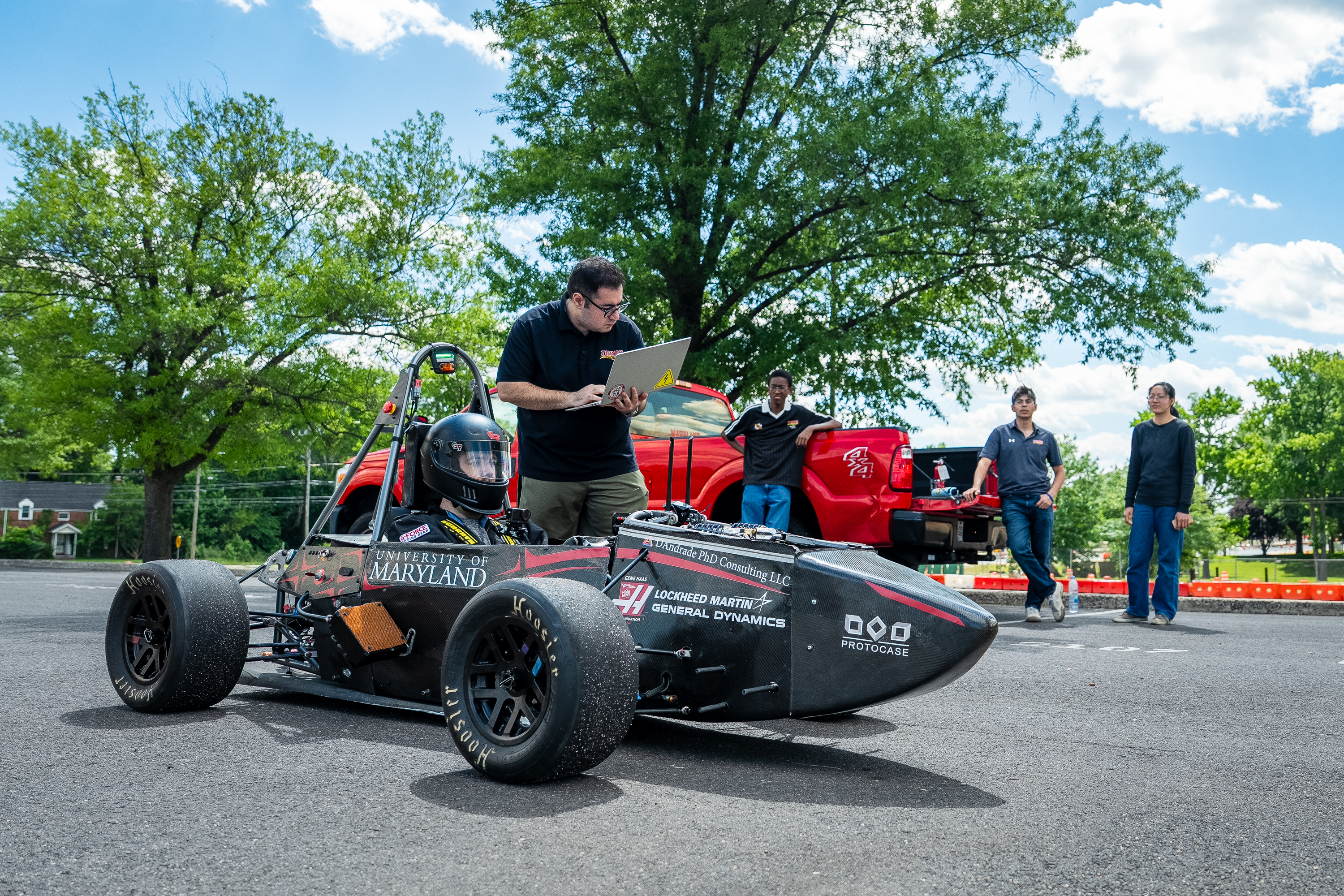 A helmeted driver sits in the car while a student beside him looks at a laptop. Other students are gathered by a red pickup truck in the background. 