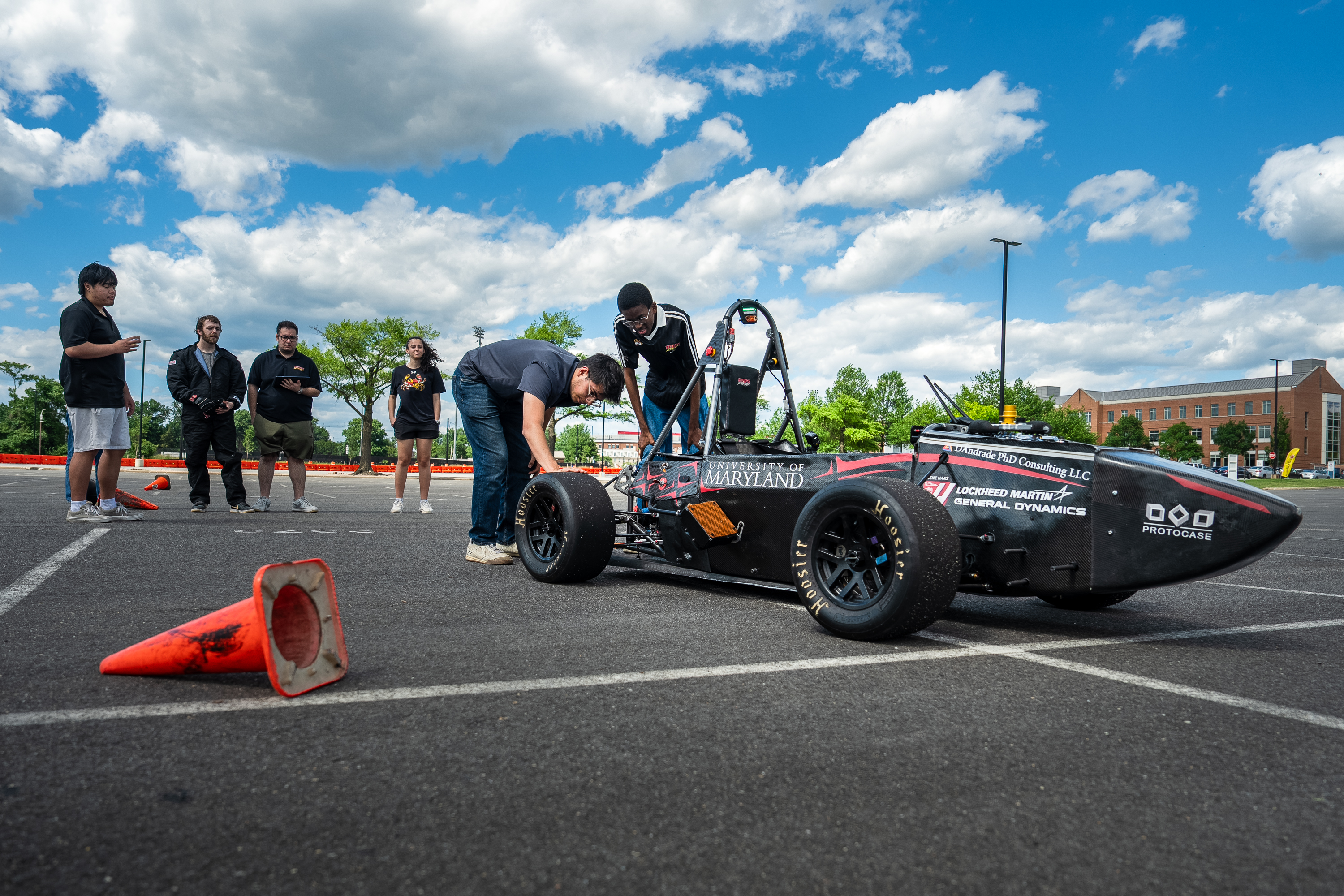 Two students inspect their car, which sits among traffic cones in a parking lot.