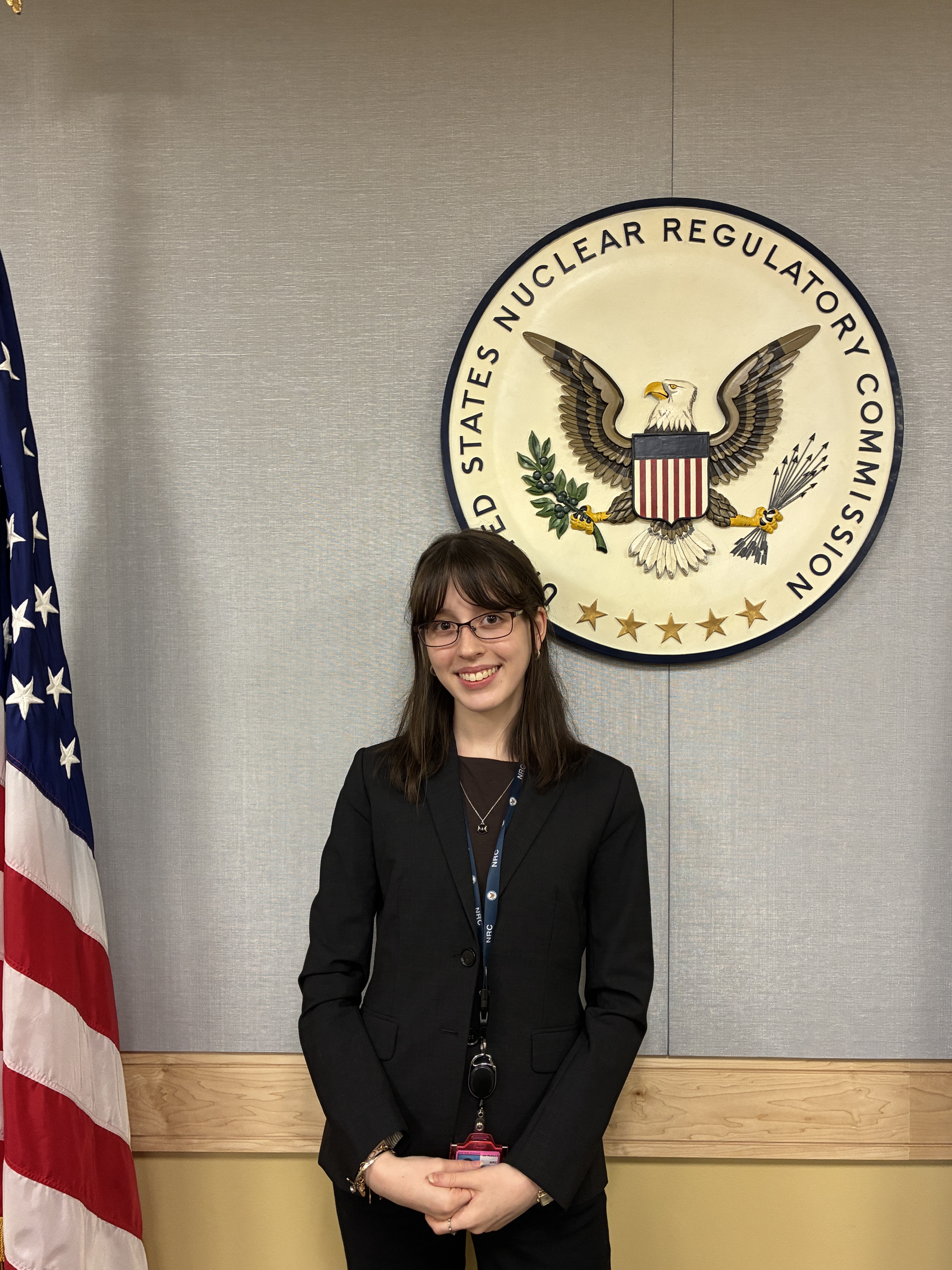 A young woman wearing a dark blazer and NRC lanyard, smiles in front of the U.S. Nuclear Regulatory Commission seal, with an American flag beside her.