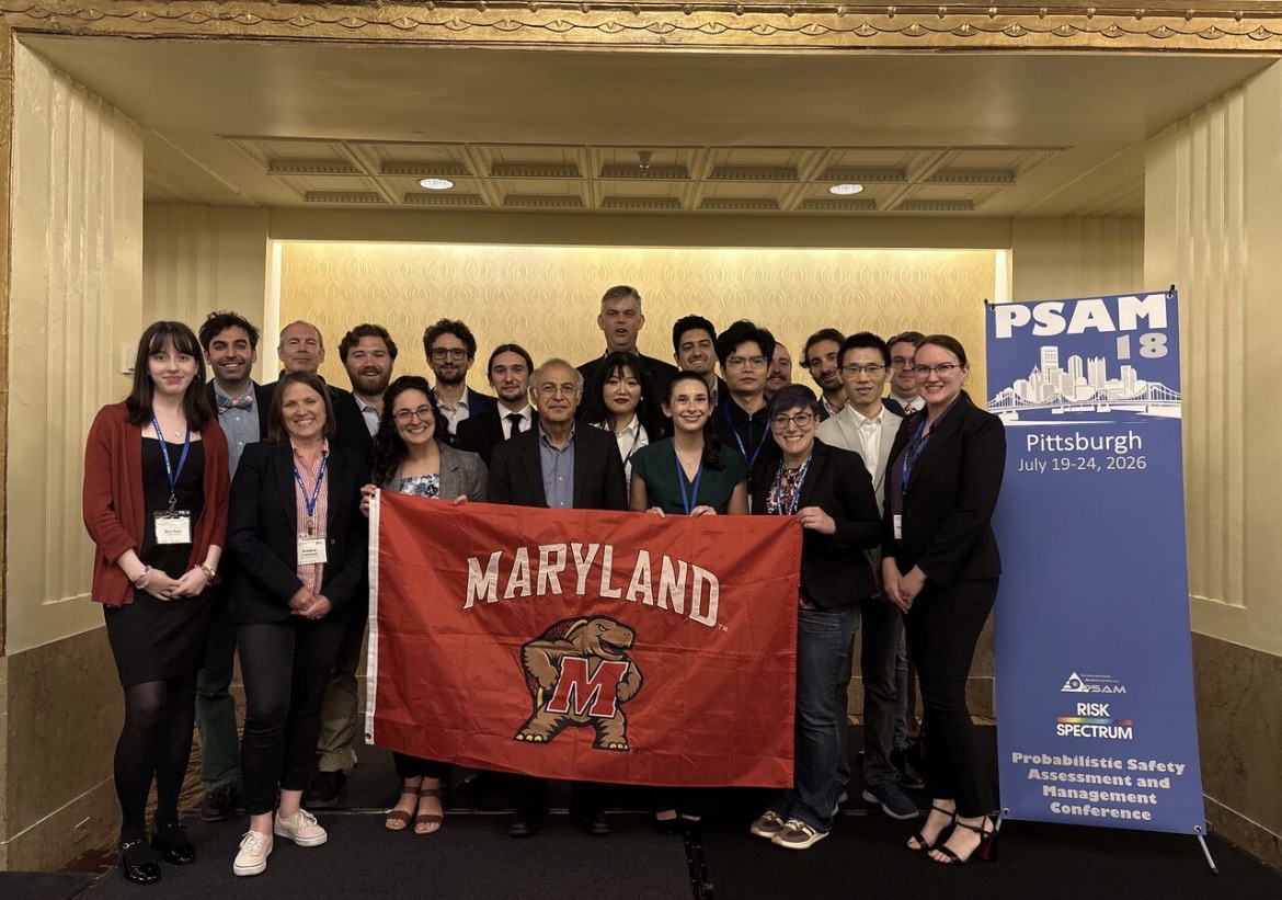 A group of about 20 people pose together holding a red Maryland flag with the Testudo logo, standing beneath a gold and beige wall at the PSAM 18 conference in Pittsburgh.