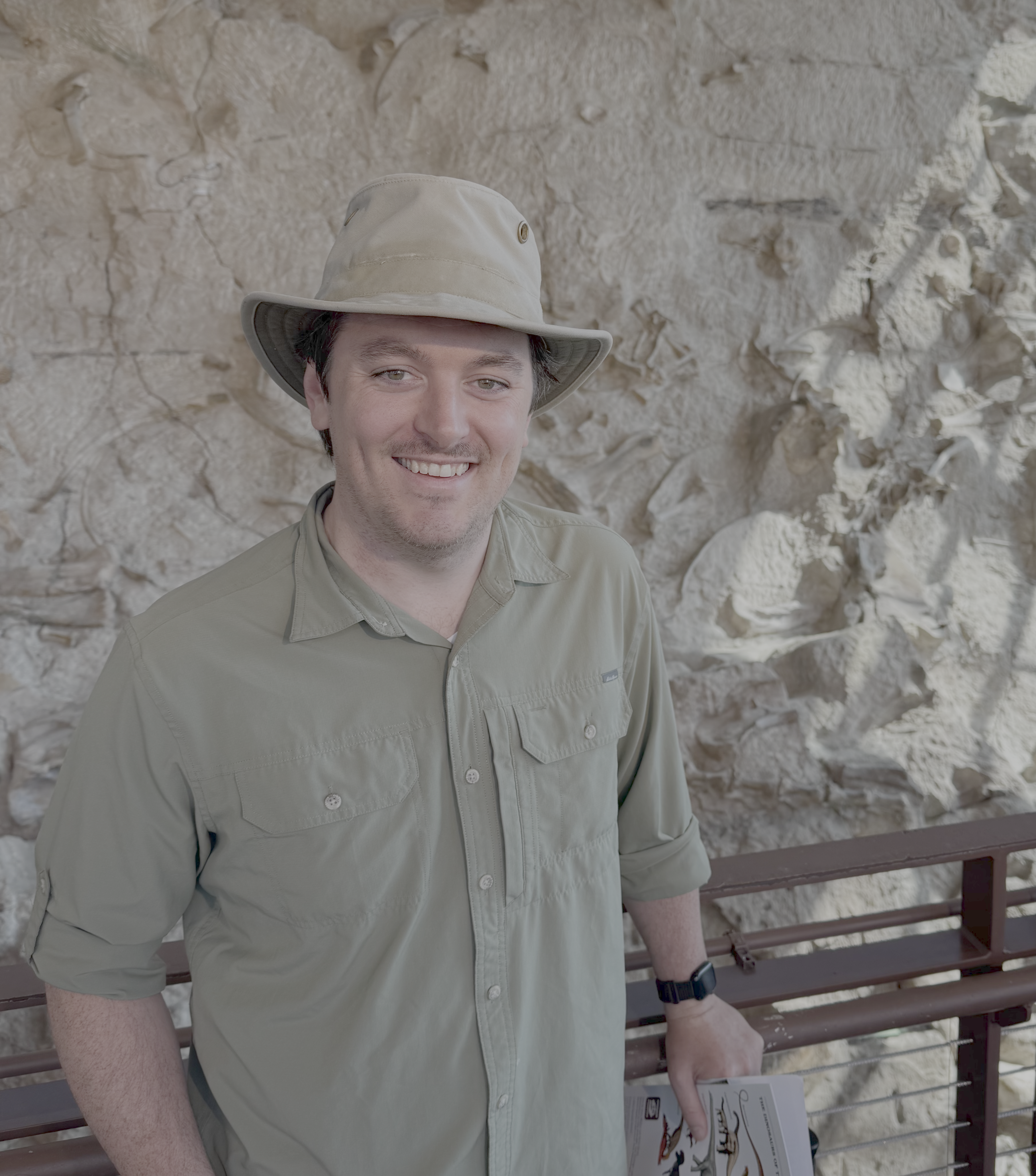 Nathan Jud standing and smiling in front of a sandstone wall in a hat and kakhi shirt.