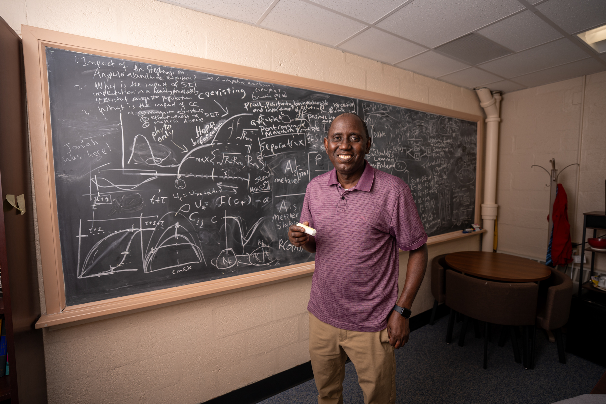 Professor Abba Gumel. wearing a red polo and khaki pants, holds a piece of chalk smiling in front of a chalkboard filled with mathematics equations and graphs. 