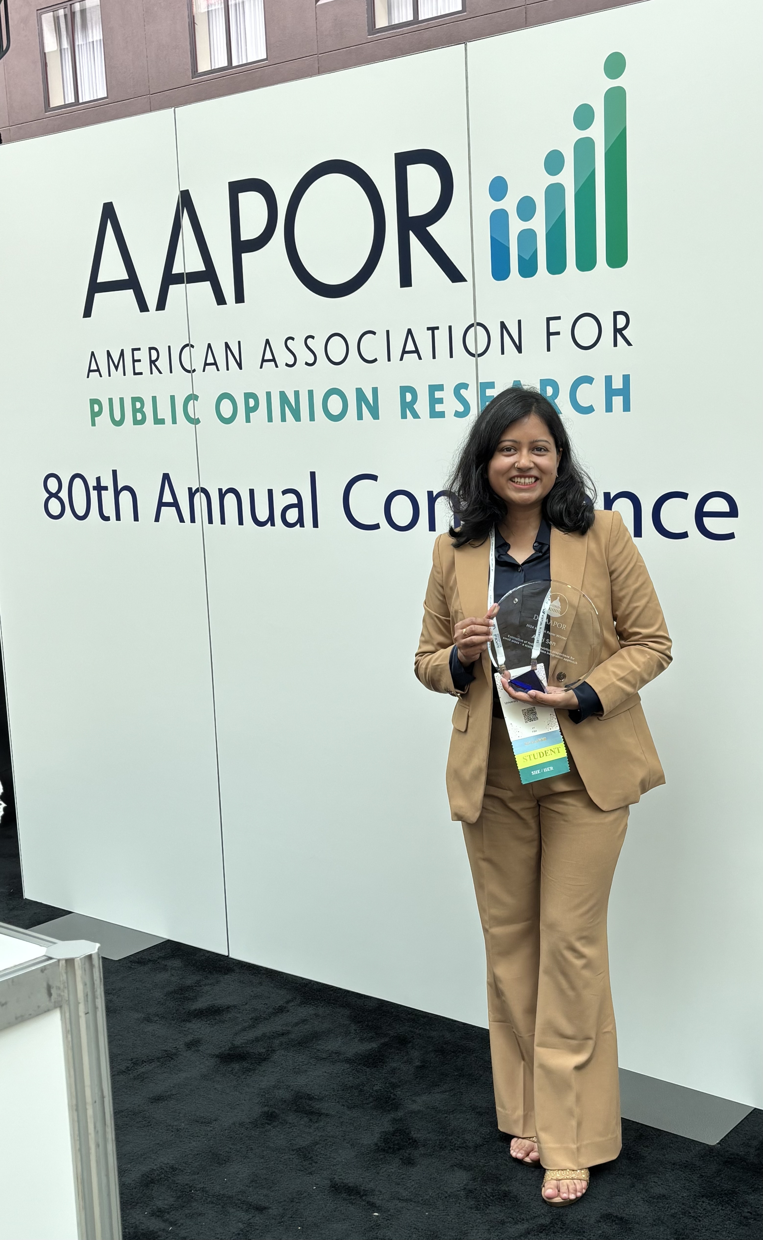 A woman in a tan suit holds a trophy in front of signage for the 80th annual conference for the American Association for Public Opinion Research (AAPOR).