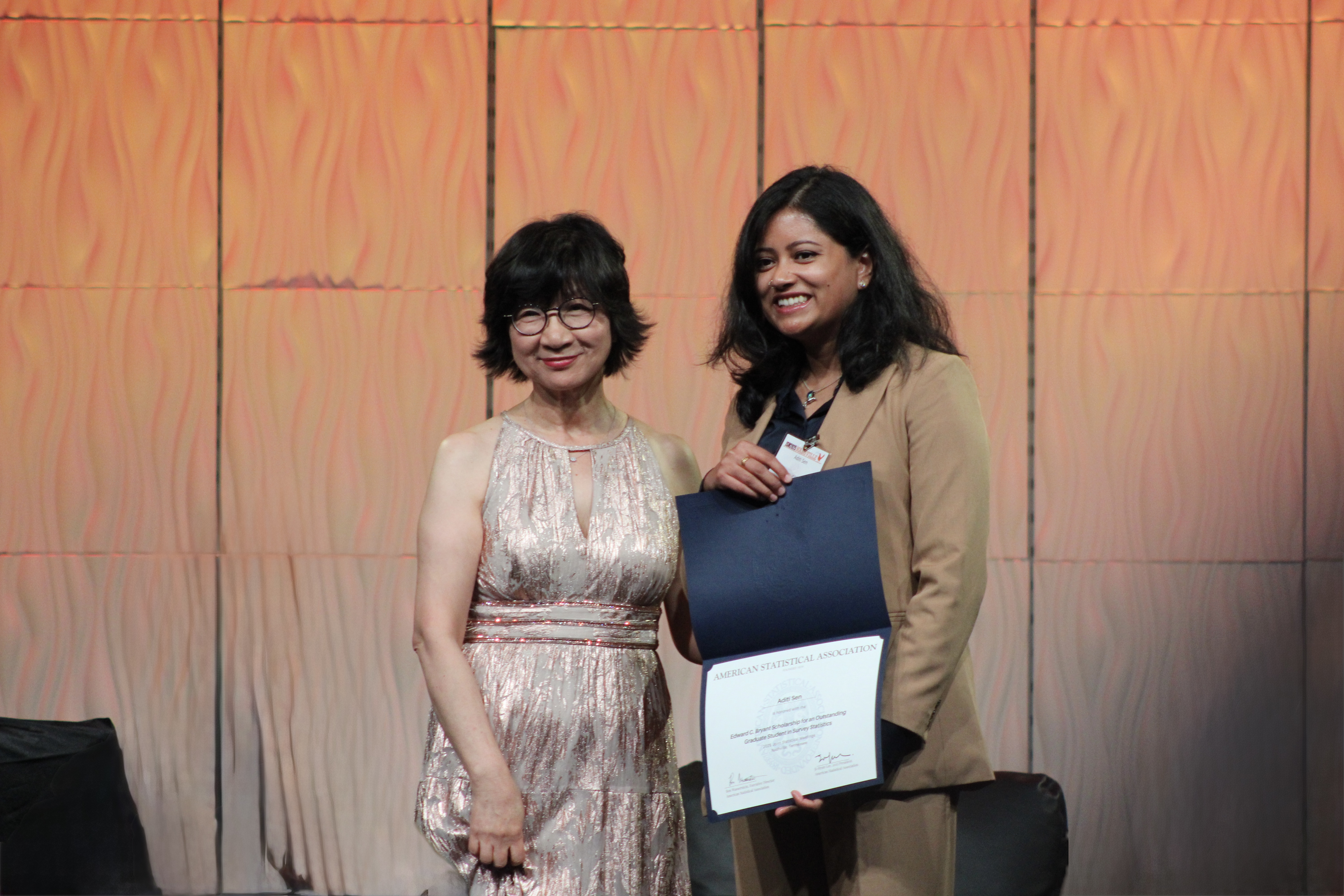 Two women standing in front of an orange backdrop, with the one on the right holding an award certificate.