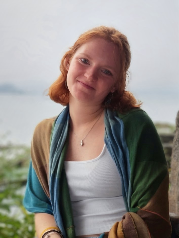 A woman stands in a white shirt and blue, green and brown coat in front of a blurred background of water.
