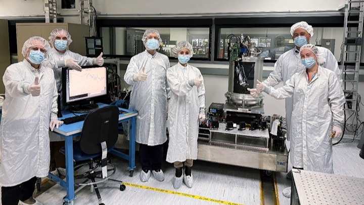 Six people in white coveralls, surgical masks, hairnets, and shoe covers standing in a laboratory setting. They are giving a thumbs-up gesture surrounded by a computer monitor displaying graph data on the left and a large, cylindrical laboratory apparatus on the right.