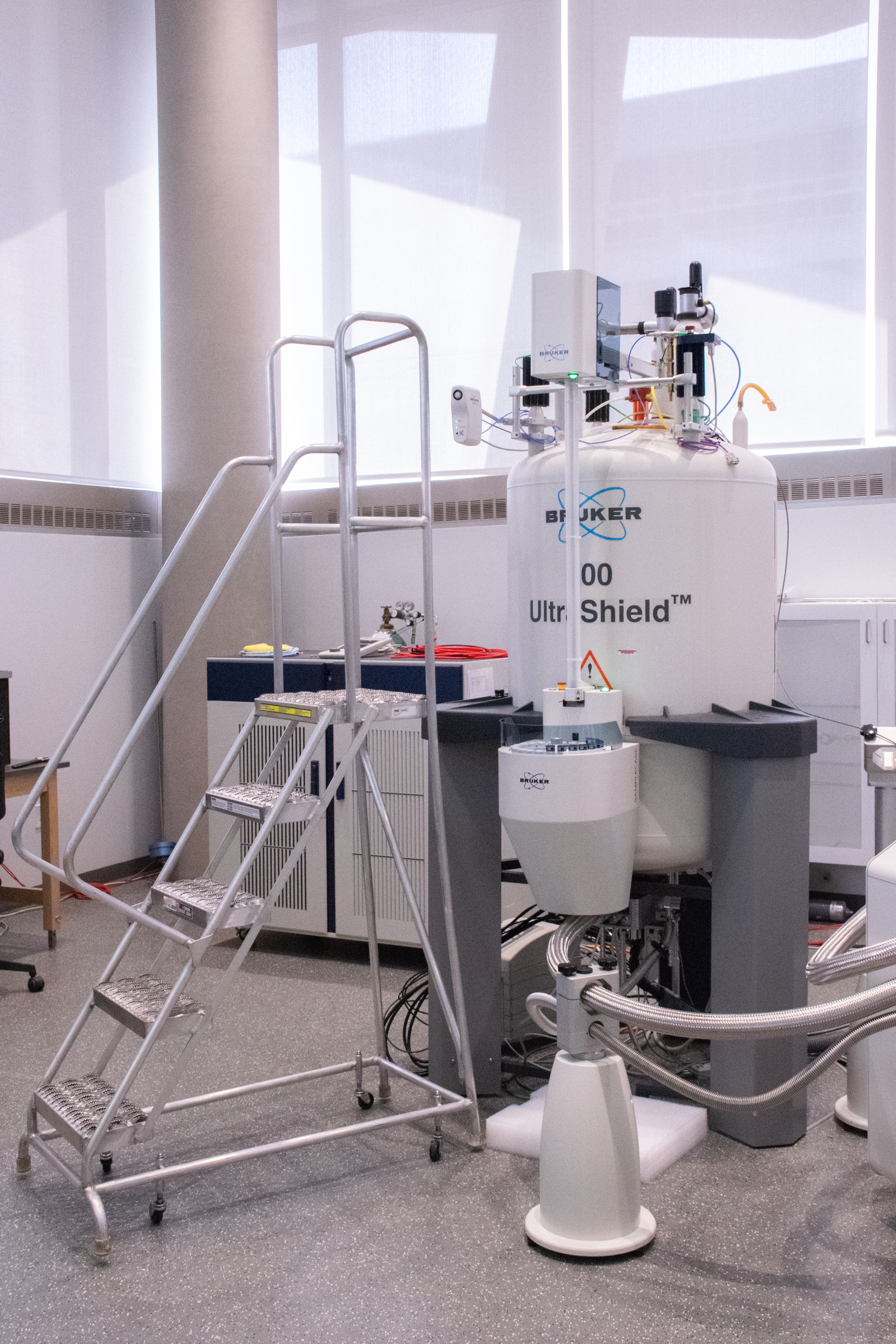 A Bruker 600 MHz NMR in a bright laboratory, surrounded by cables and equipment, with a metal stair platform positioned next to it.