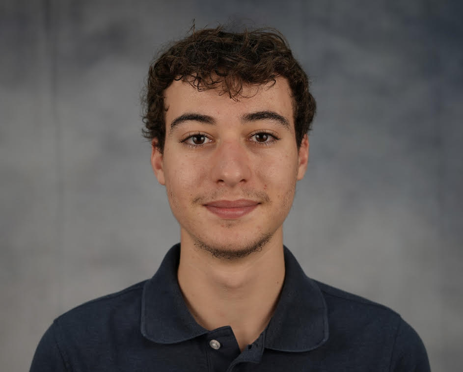 Headshot of a man with curly hair wearing a navy blue polo shirt in front of a mottled gray backdrop. 