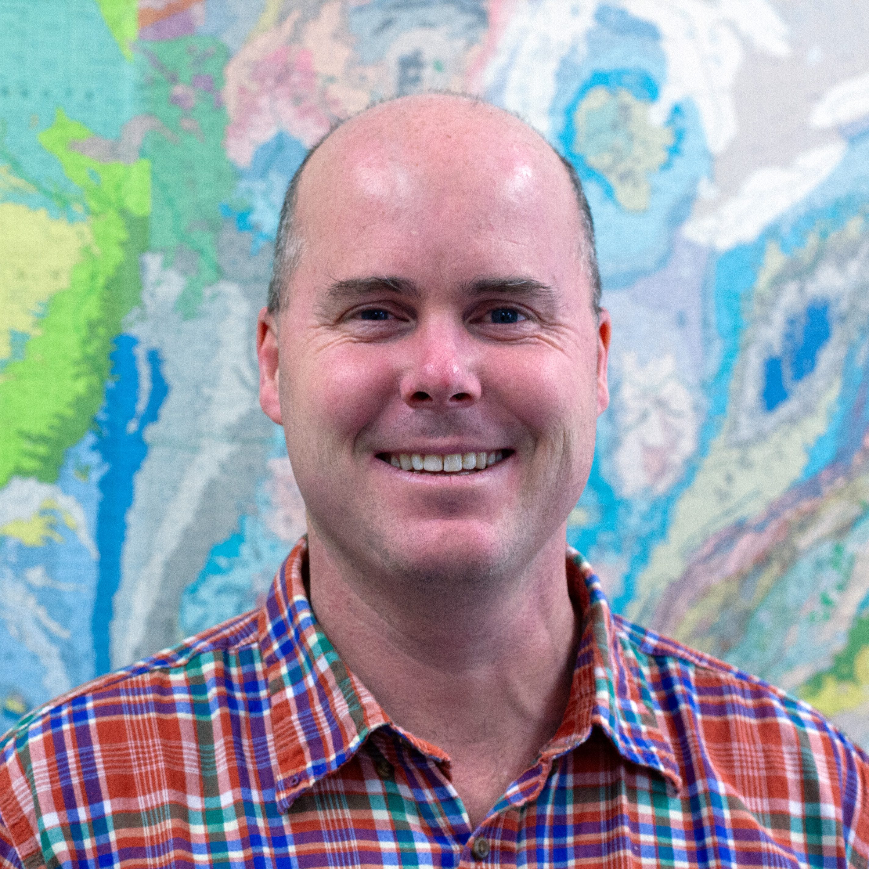 A man in a plaid shirt stands in front of a colorful map.