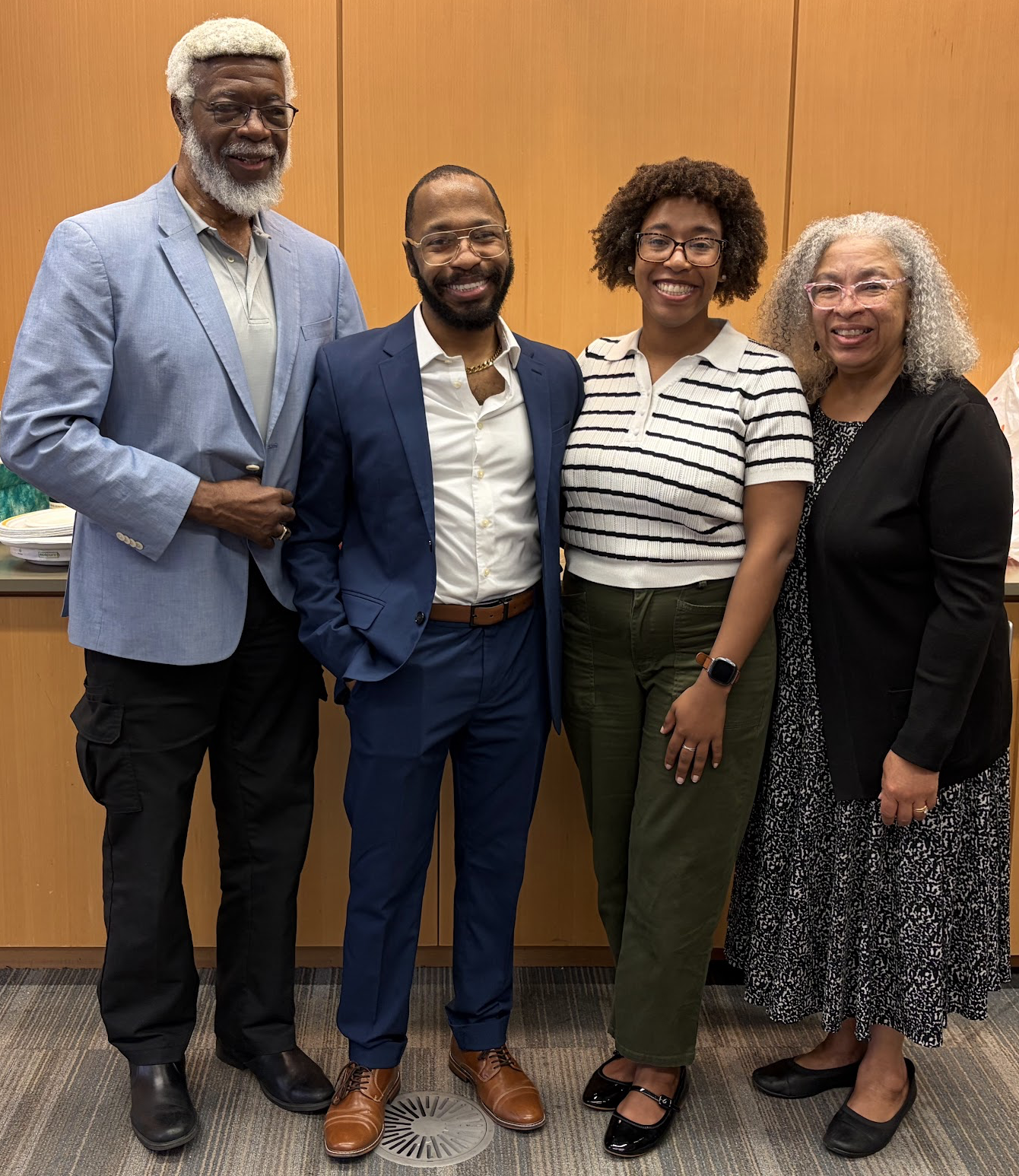 Four adults stand together smiling in front of a light wood-paneled wall. From left to right: a tall man with white hair and beard wearing a light blue blazer and black pants; a man in a navy blue suit with brown shoes; a woman wearing glasses, a white striped short-sleeve sweater, and olive green pants; and a woman with curly gray hair wearing glasses, a black cardigan, and a patterned black-and-white dress. 