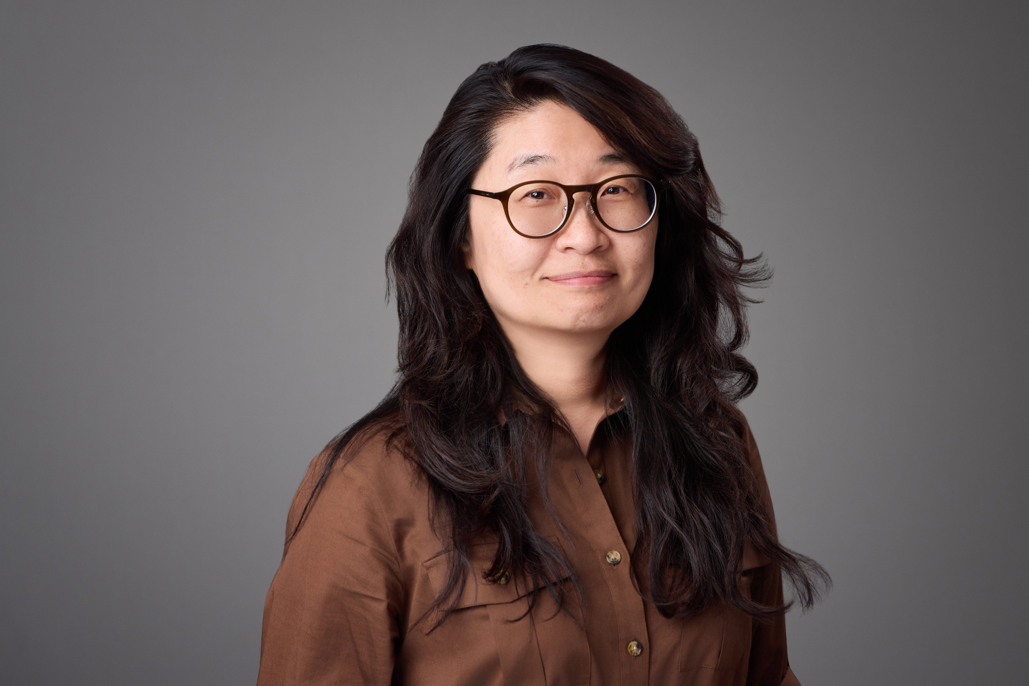 Portrait of a woman wearing glasses and a brown button-down shirt against a neutral grey background.