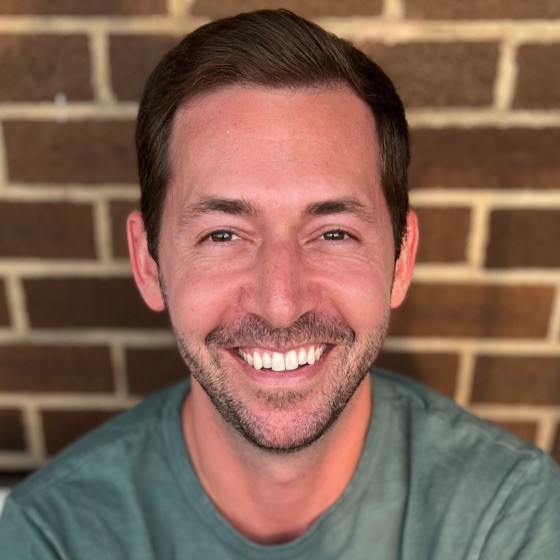 A man with brown hair and facial hair smiles wearing a dark green shirt in front of a brick wall. 