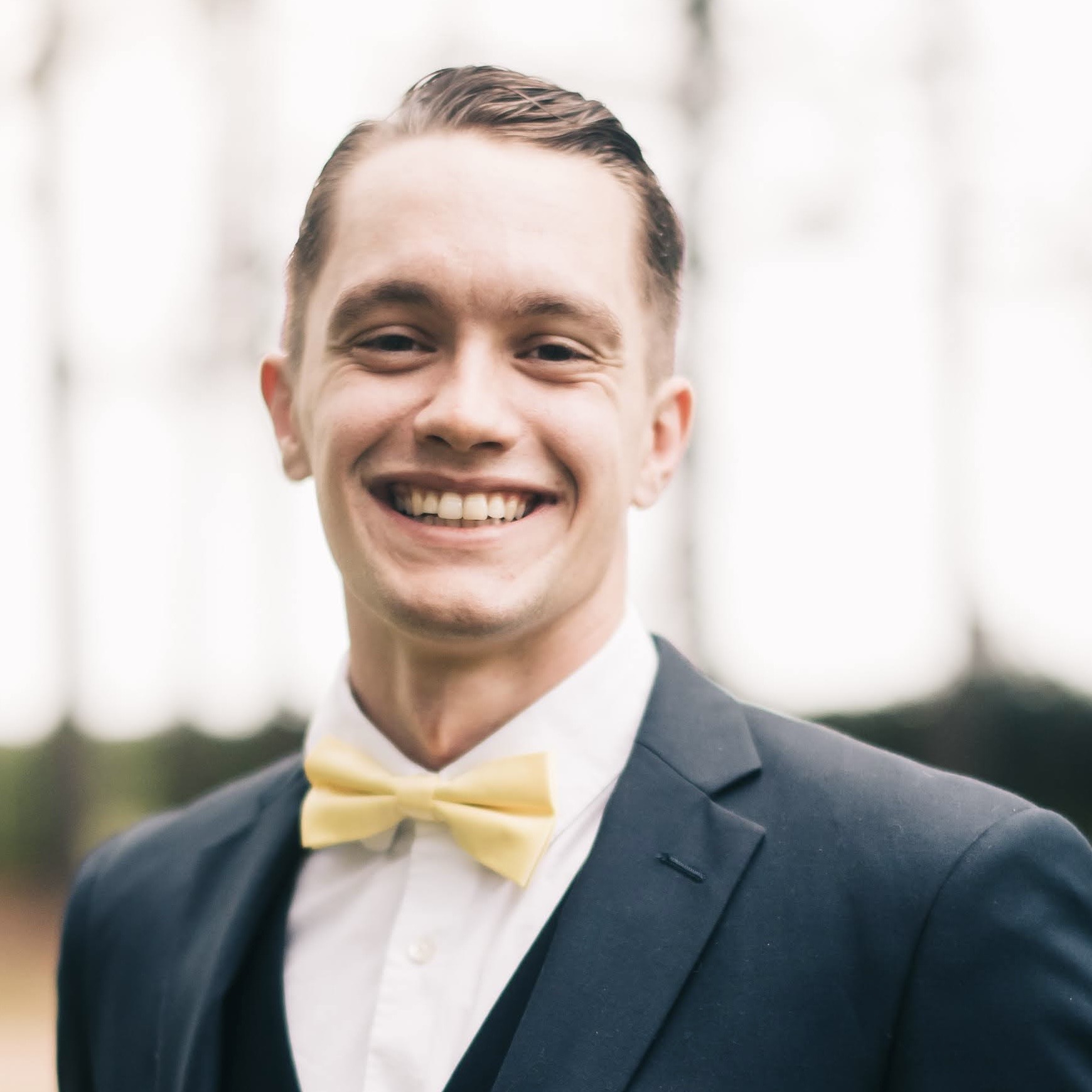 A man smiles wearing a black suit and yellow bow tie.