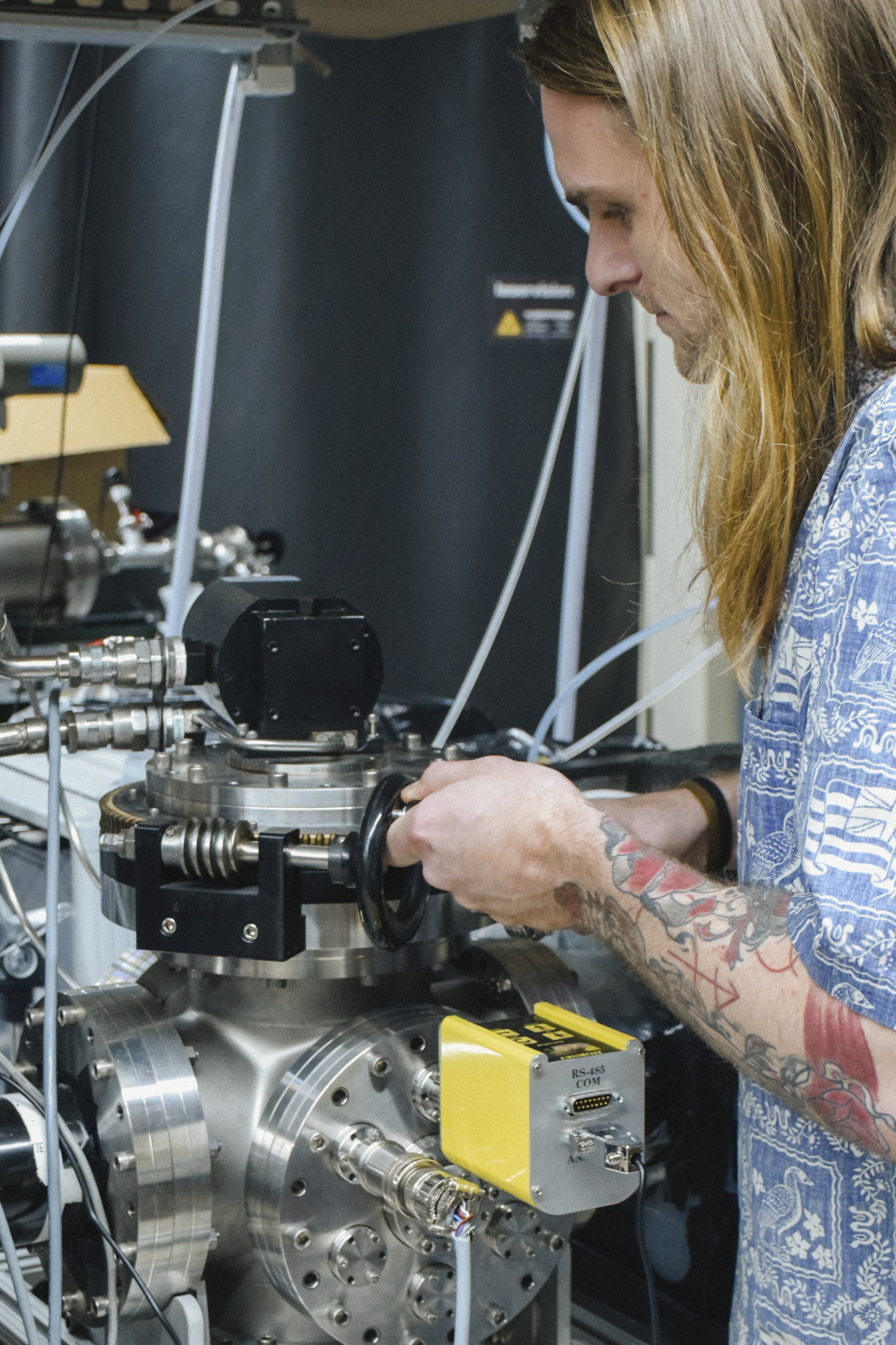 A man with long, blonde hair in a blue short-sleeved shirt operates a silver piece of scientific equipment.