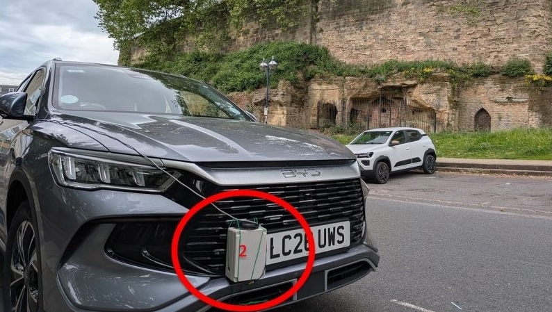 Close-up of a dark grey BYD vehicle parked on a road next to an old stone wall structure. Encircled in red on the front bumper is a small grey plastic box with a red "2" written on it, secured to the front grill next to the UK license plate.