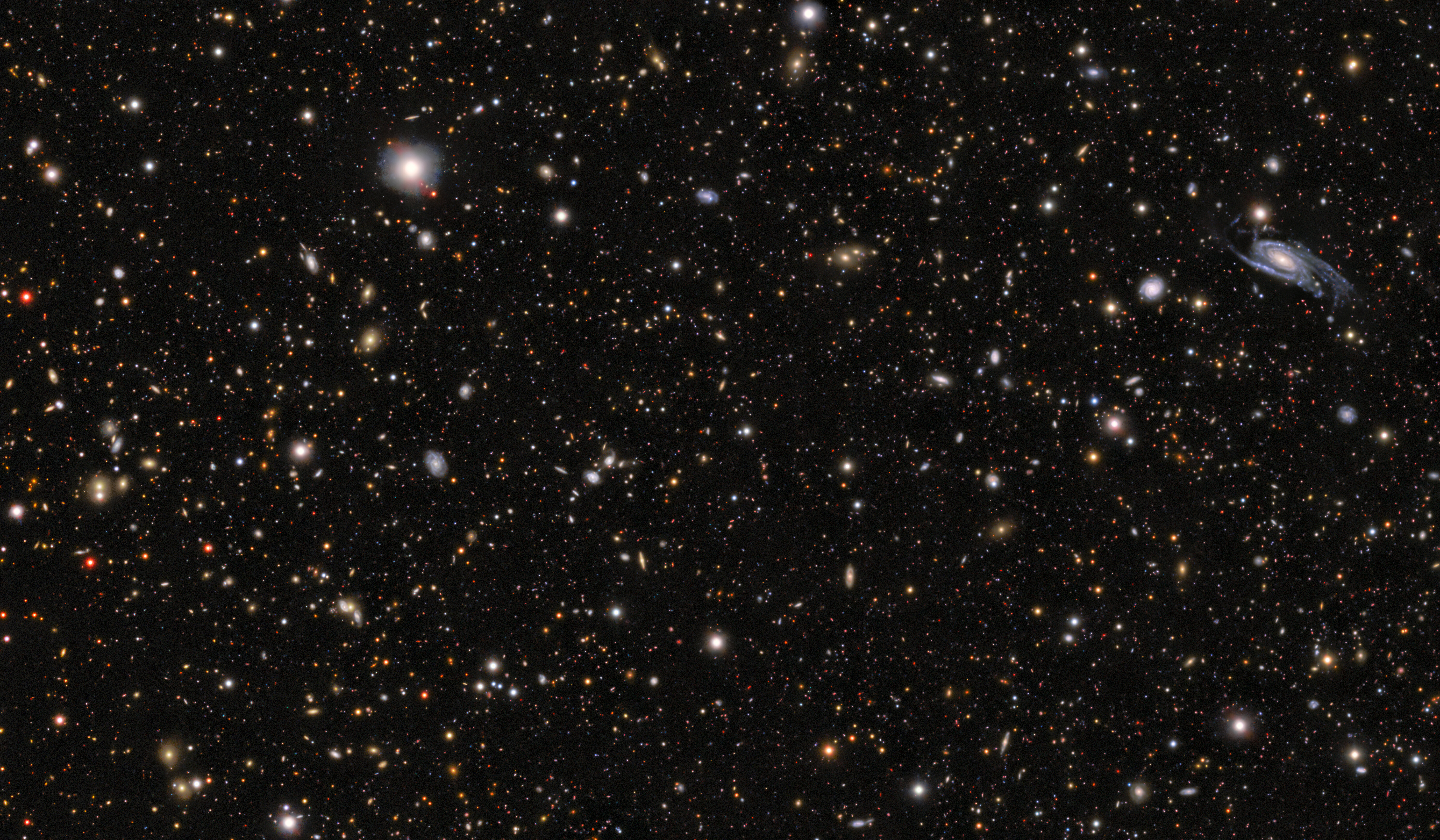 Telescope image depicting a black sky with hundreds of red, yellow, white and blue dots, plus a blue galaxy in the top-right of the image.