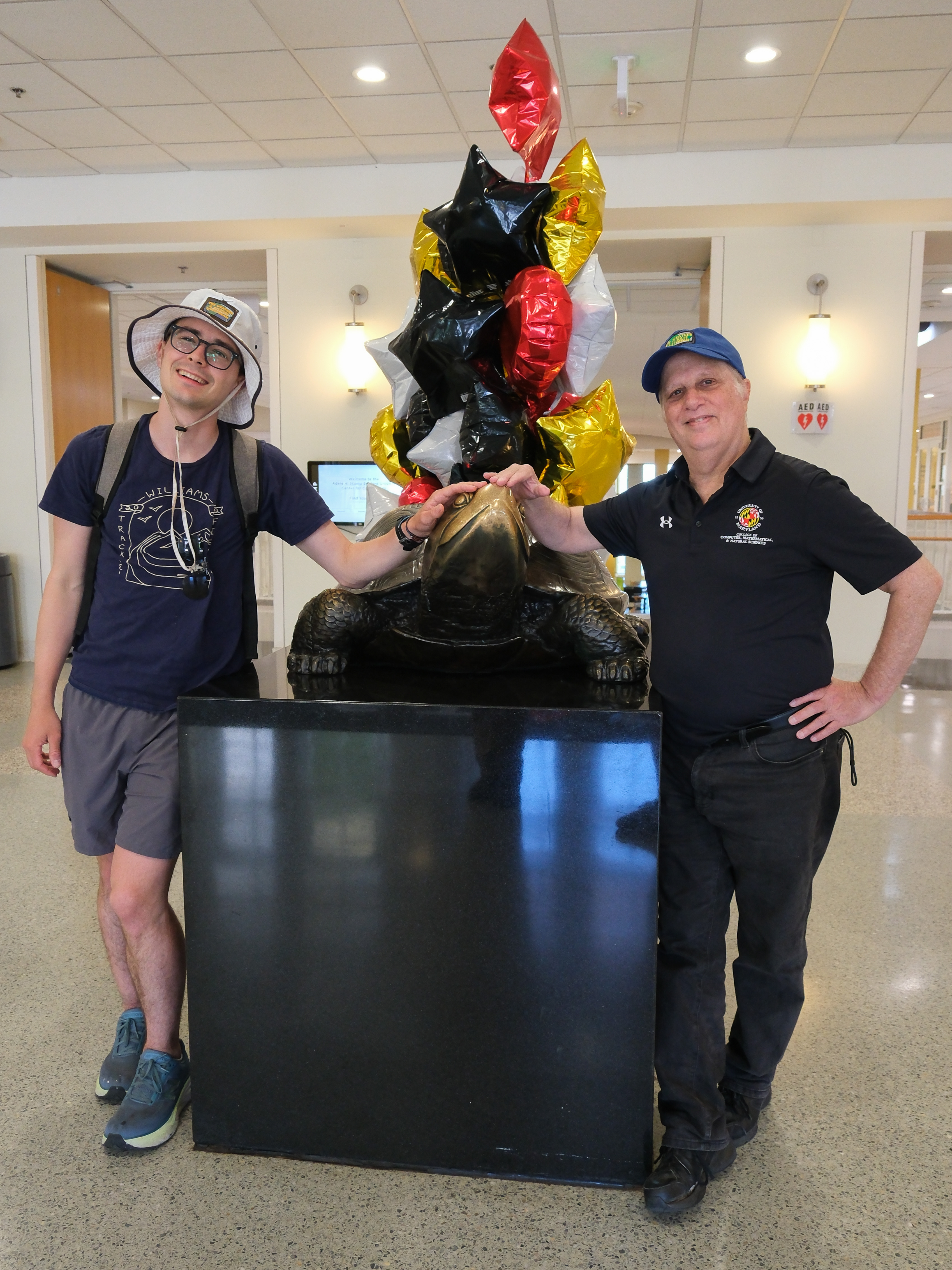Two men stand inside touching the nose of a statue of a tortoise. The one on the left wears gray shorts, a navy t-shirt, and a light gray bucket hat. The one on the right wears black jeans, a black University of Maryland promo, and a blue baseball cap.