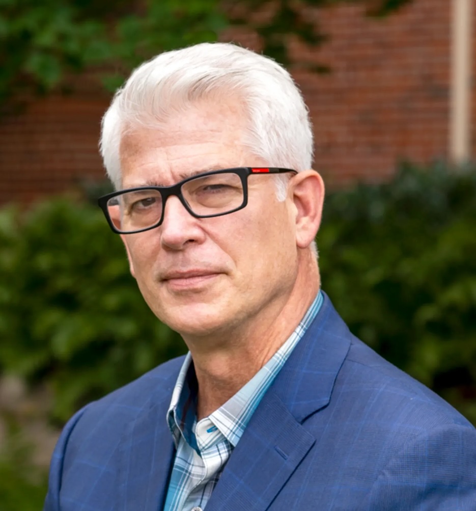 A man in black glasses and a blue suit over a blue plaid shirt stands in front of blurred greenery and bricks.