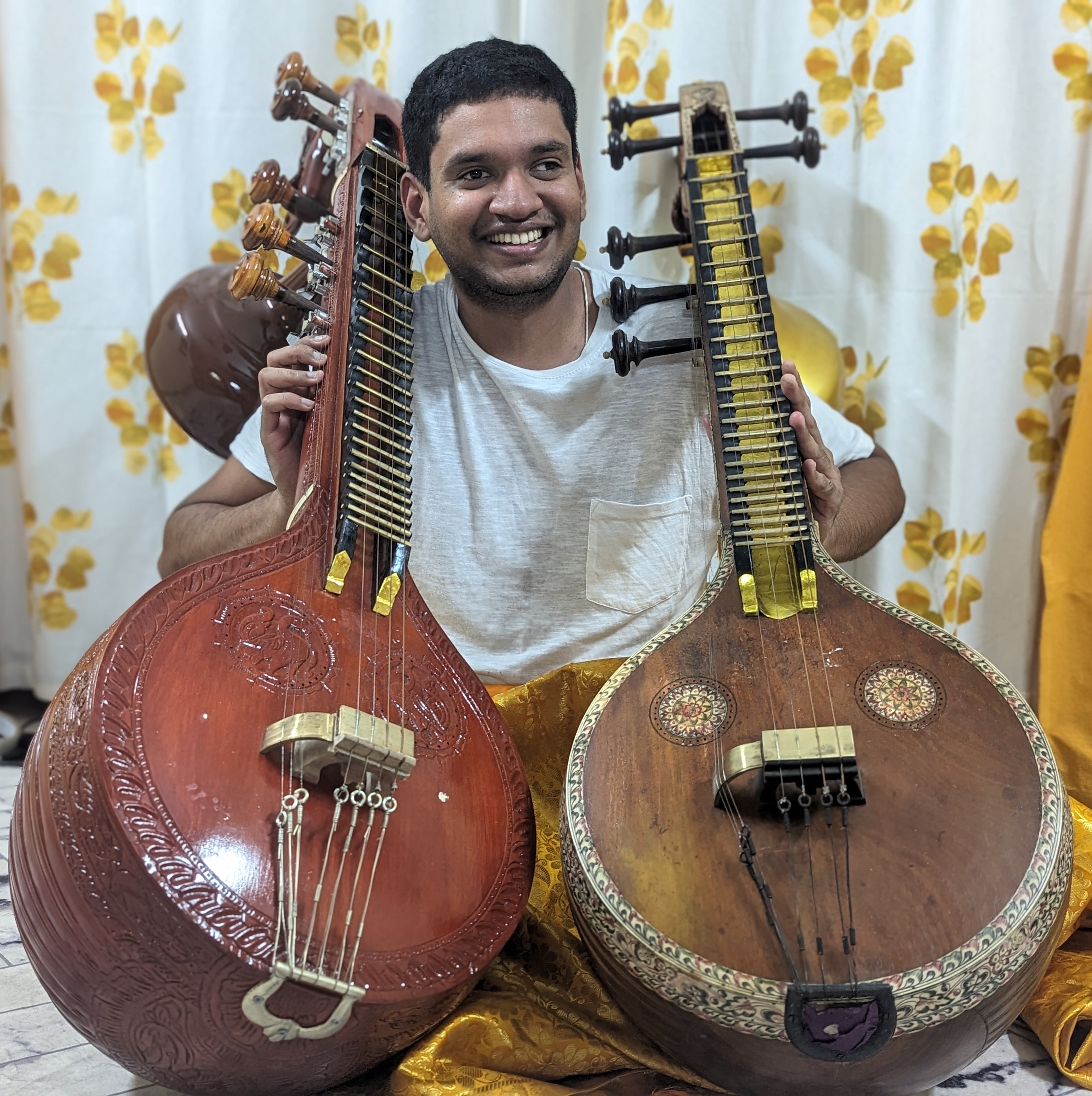 Srivatsav Kunnawalkam Elayavalli holding two veenas sitting in front of a white and gold curtain.