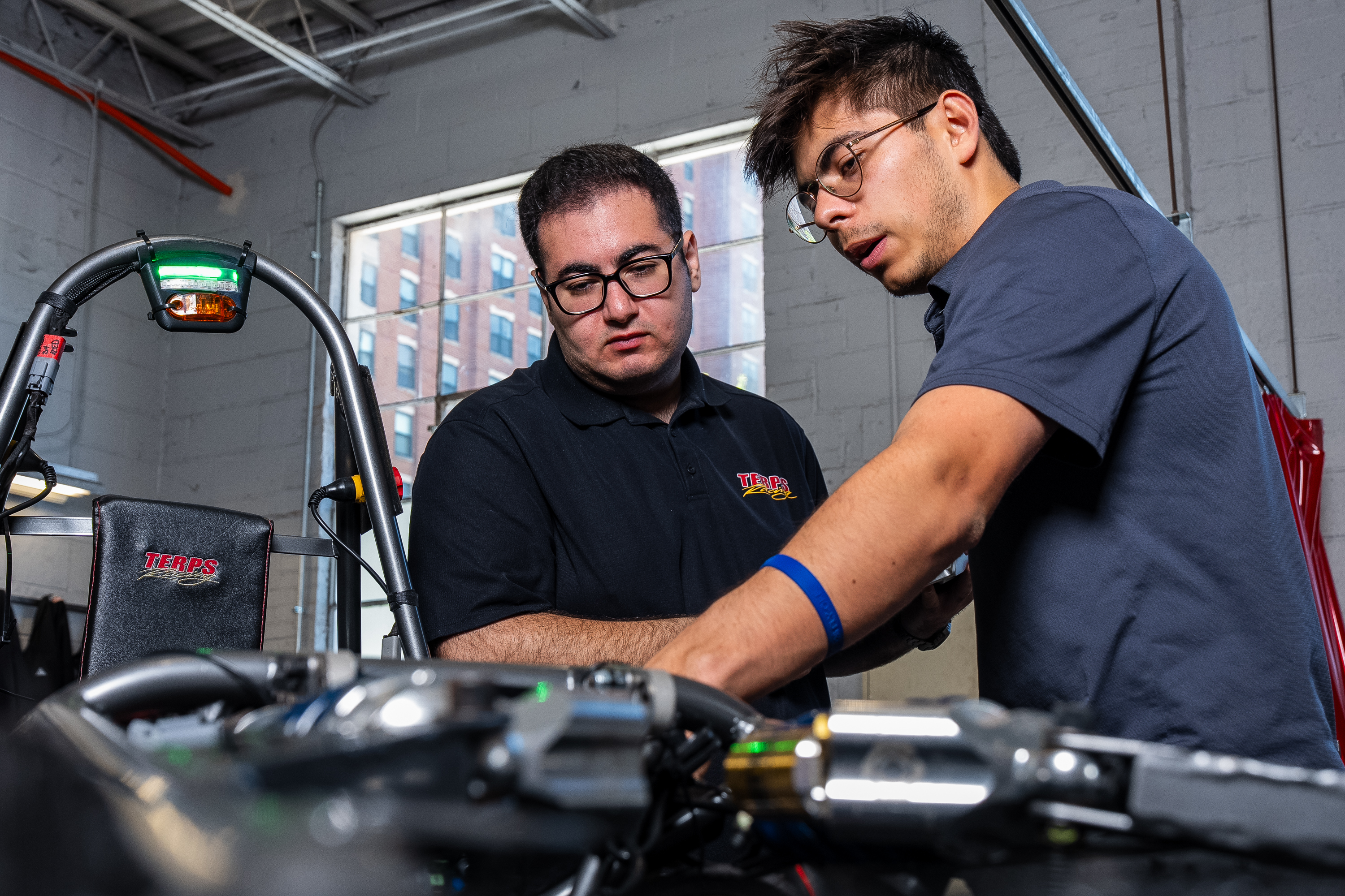 Two young men wearing polos point toward the inside of a black race car in a dimly lit garage.
