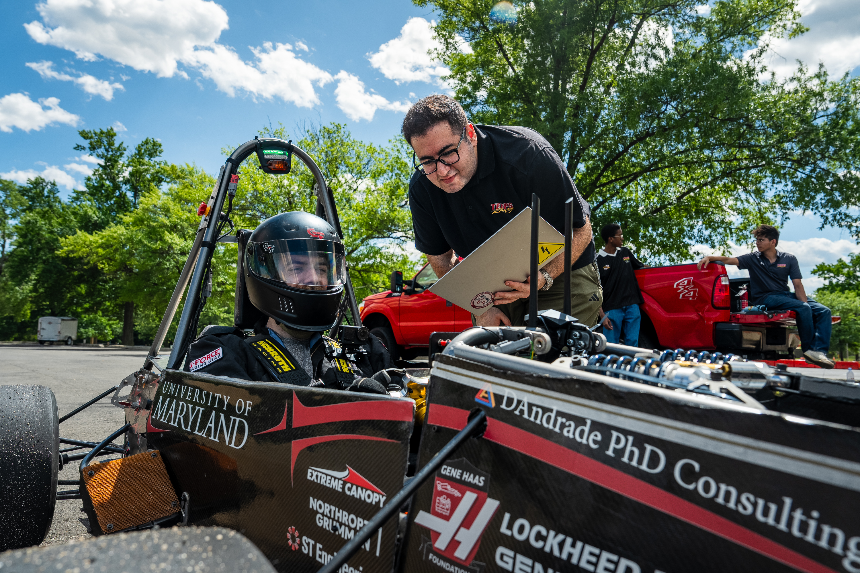 A person in a black racing suit and helmet sits inside a black race car with the words "University of Maryland" and various corporate sponsors, while a man holding a laptop speaks to him. 