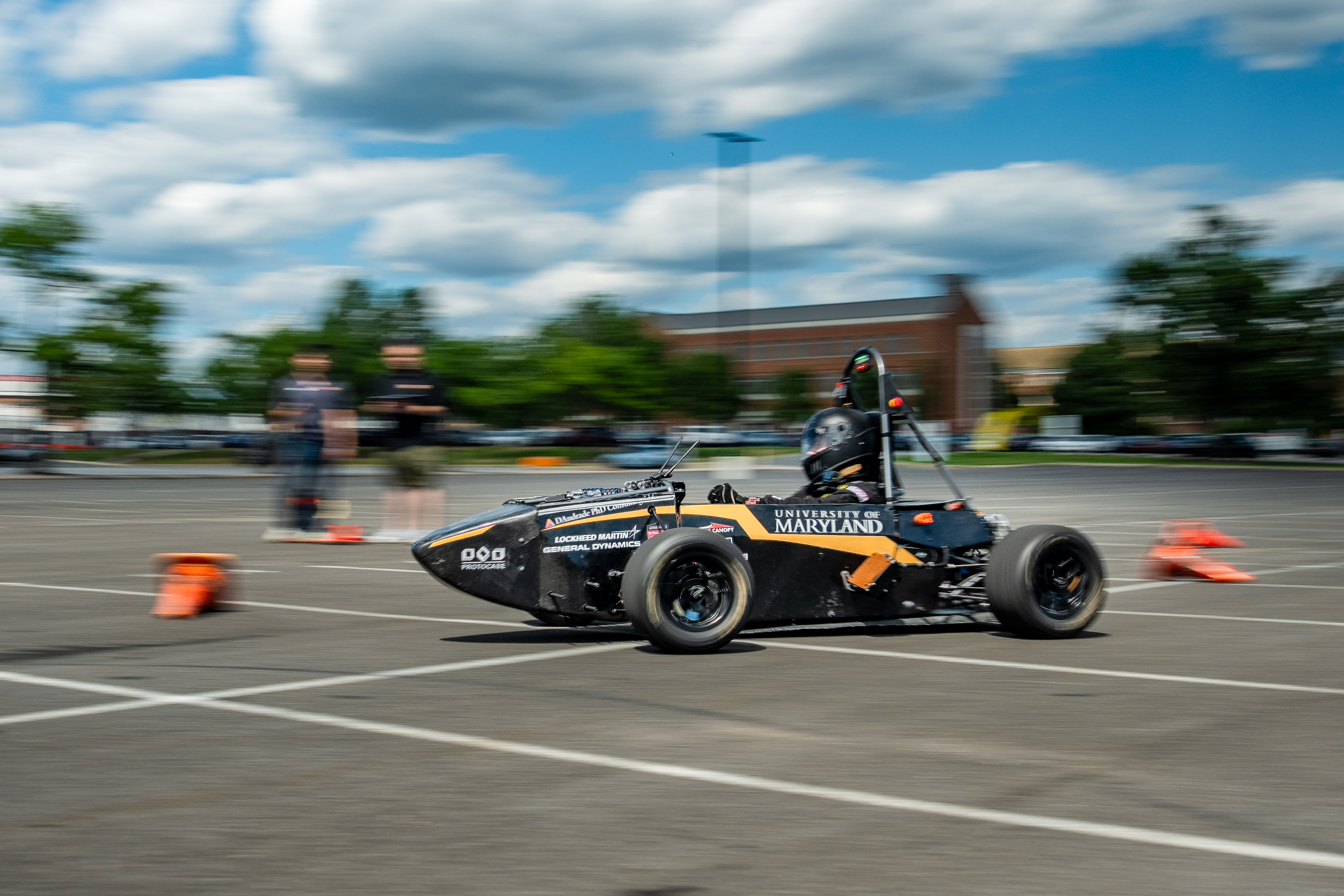 A black racecar with the text "University of Maryland" moves along a blurry background of a blue, cloudy sky and green trees.