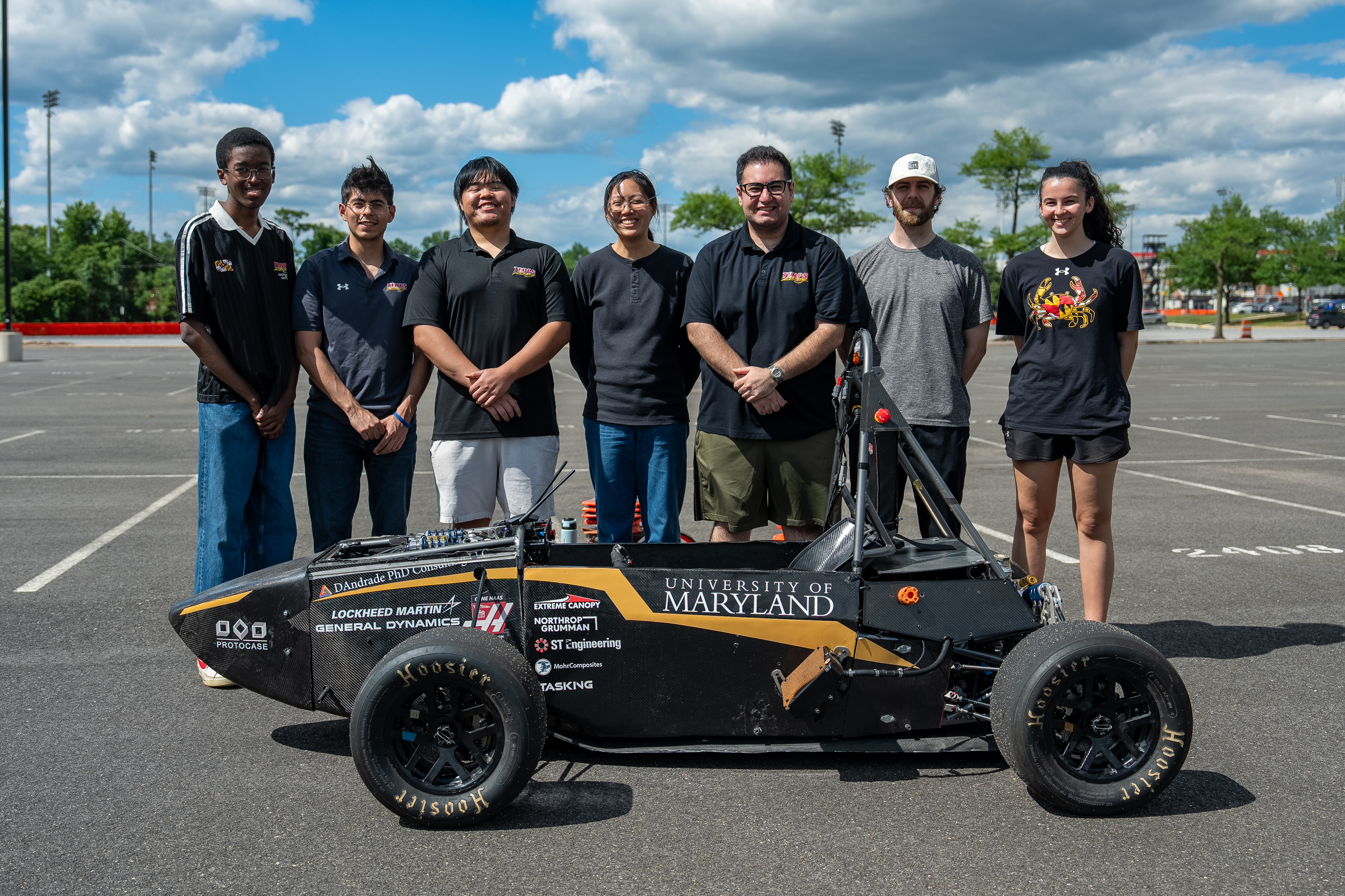 Seven students stand behind a small black race car under a blue, cloudy sky.