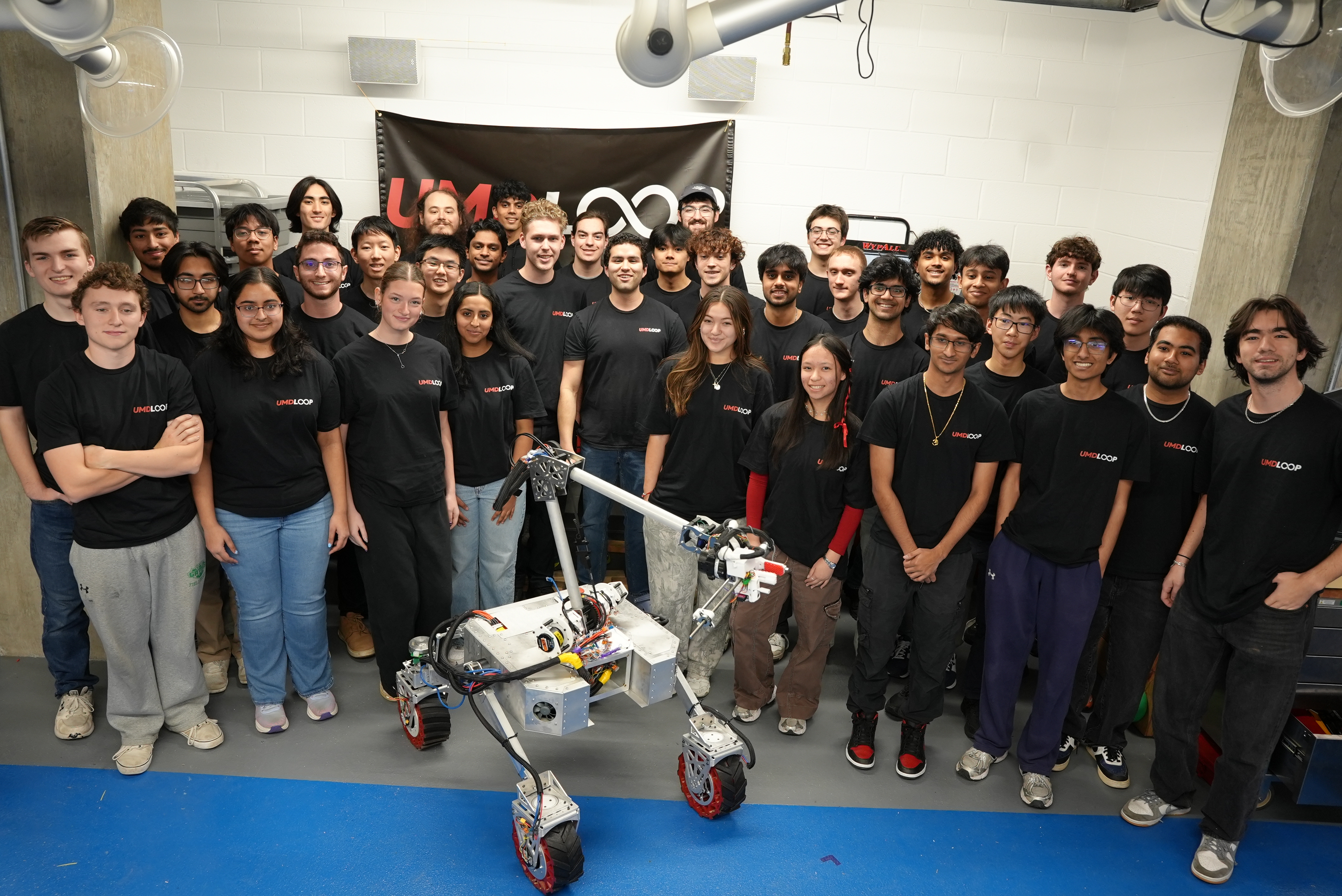 A large group of students wearing matching black UMD Loop shirts pose indoors behind a four-wheeled robotic rover with a mounted mechanical arm, in a lab space with a UMD Loop banner on the wall behind them.