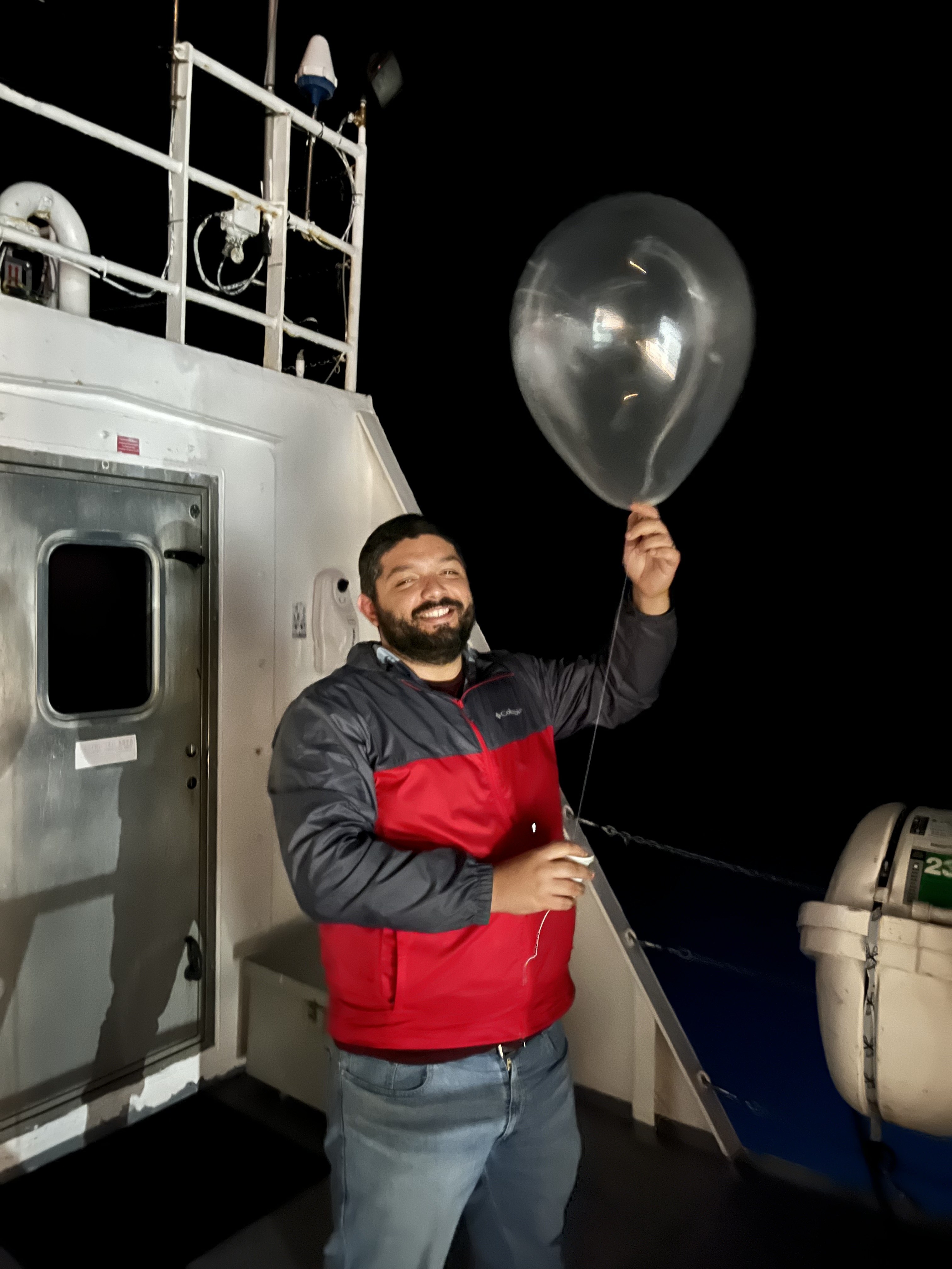 A smiling man in a red and black rain coat holds a weather balloon on a ship at night.