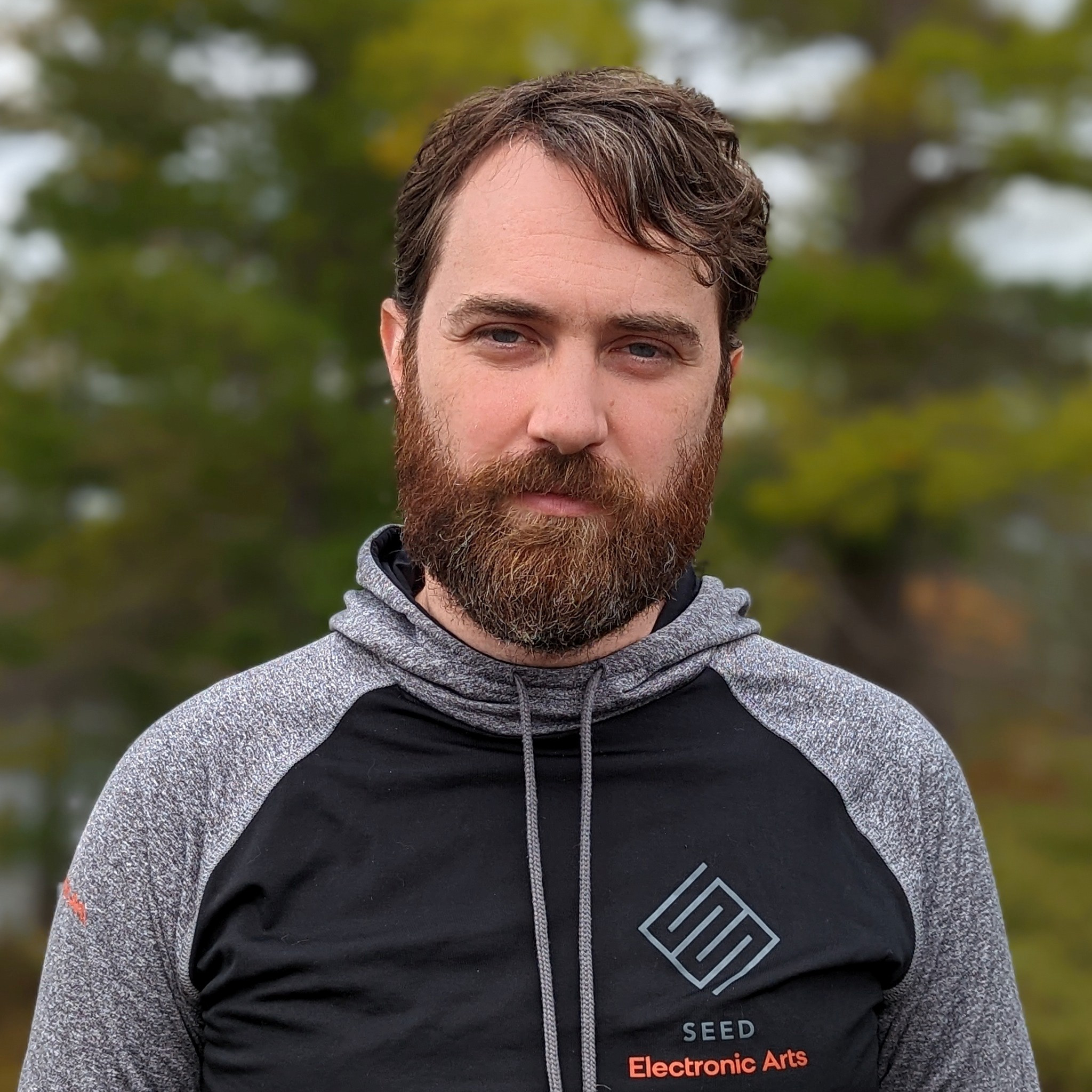 A bearded, brunette man wears a black and gray hoodie that says "SEED Electronic Arts" standing in front of a blurred background of trees.