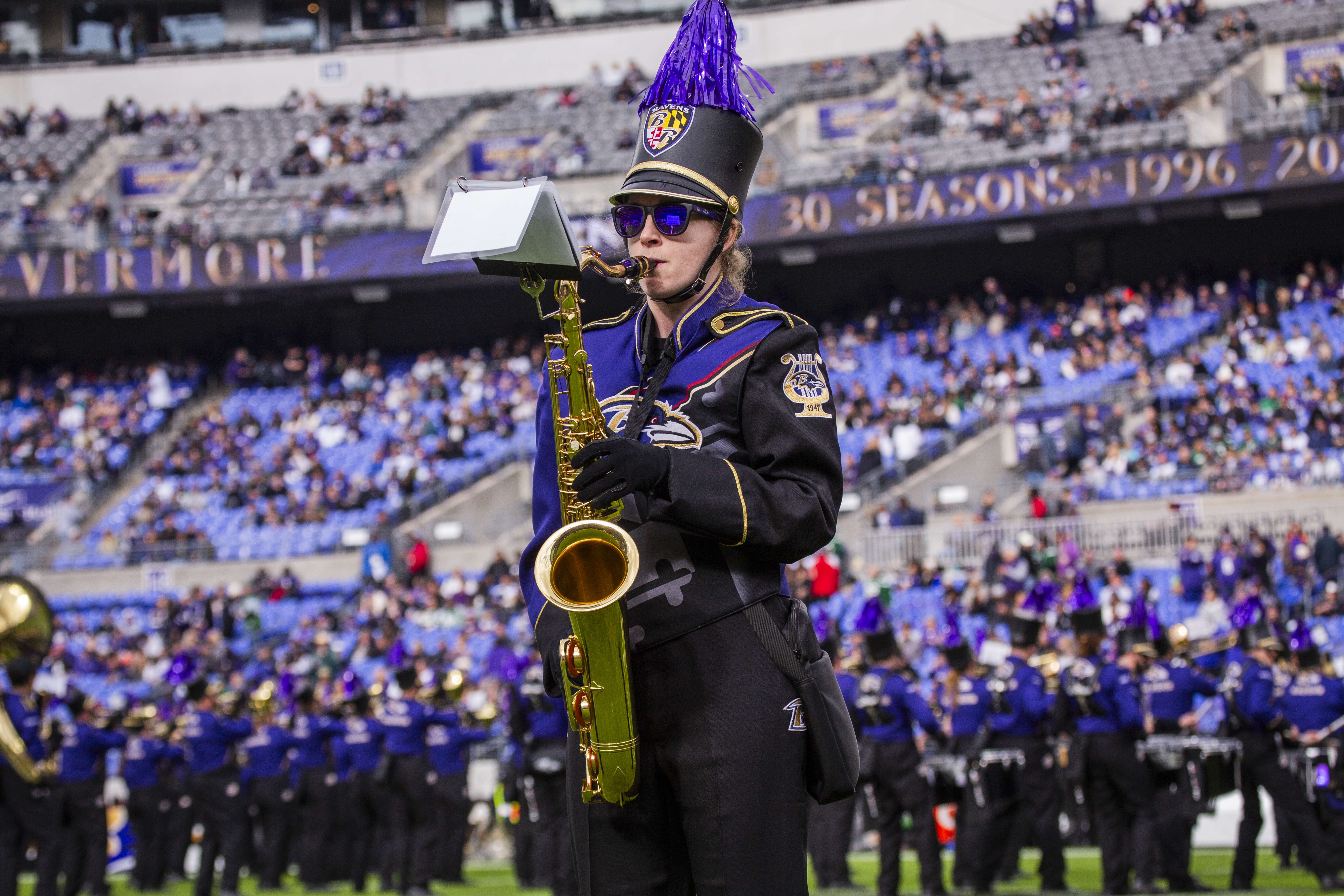 A member of a marching band stands on a field holding a tenor saxophone, wearing a detailed purple and black uniform with gold trim, a black hat topped with a tall purple feather plume, and dark sunglasses. The blurred background shows a crowded stadium seating area under a "30 SEASONS" sign and other band members lined up on the field.