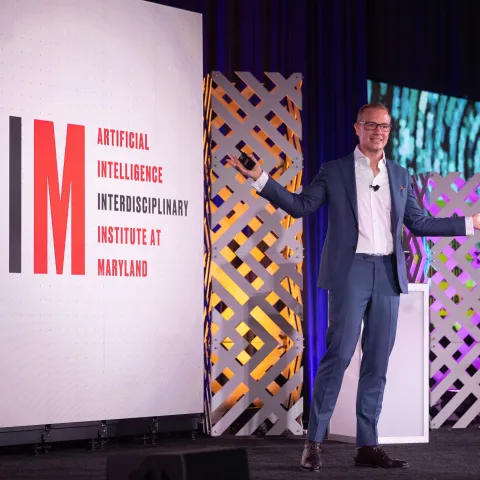 Hal Daume wearing a suit speaking on stage, with a large AIM sign to the left