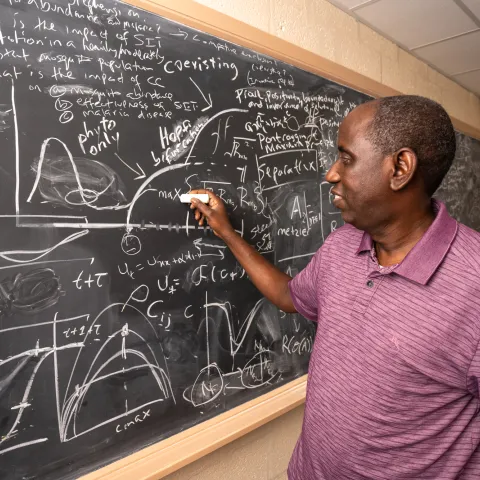 Man holding a piece of chalk and writing on a blackboard filled with graphs and equations
