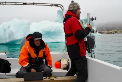 Cy Keener (seated on a boat at an iceberg in Greenland while Lars Smedsrud holds device equipped with sonar technology