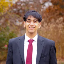 A male college-aged student wearing a black suit with a white dress shirt and red tie stands in front of a blurred background of autumnal-colored foliage.
