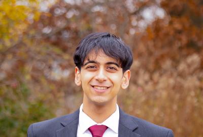 A male college-aged student wearing a black suit with a white dress shirt and red tie stands in front of a blurred background of autumnal-colored foliage.