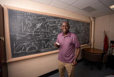 Professor Abba Gumel. wearing a red polo and khaki pants, holds a piece of chalk smiling in front of a chalkboard filled with mathematics equations and graphs. 